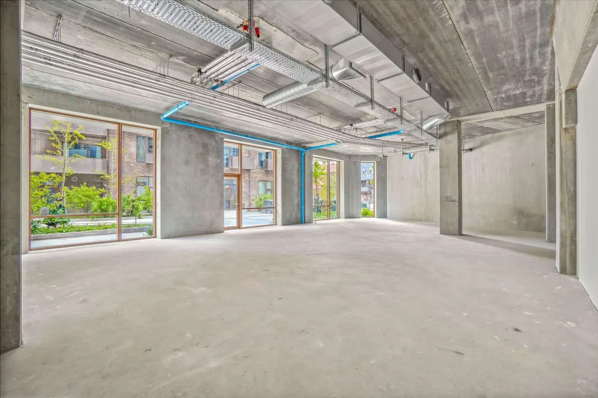 Lejemål med store vinduer og højloft i København. Betongulv og ventiler i loftet. Udsigt over grønne områder og bygninger