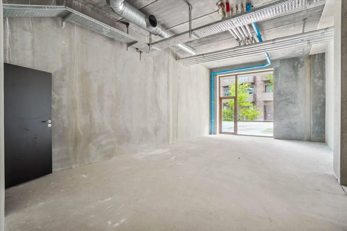 Store, højloftede rum med vinduesparti og afkrydsningsfelter på loftet. Betonvægge og gulv samt et sort dørblad til venstre.