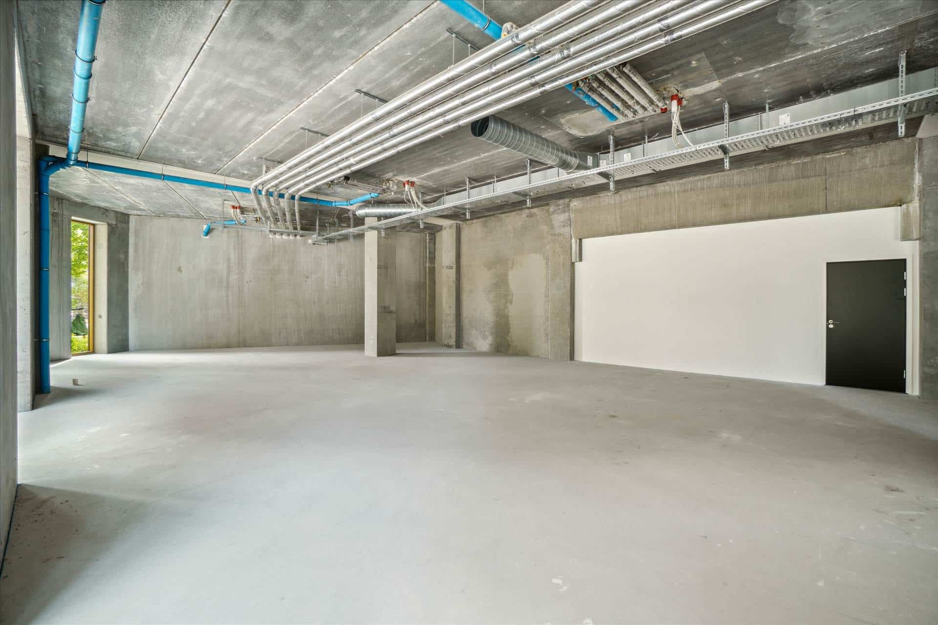 En tom lejlighed med højloft, tom beton og vinduer i bagvæggen, samt rørmærker på loftet.