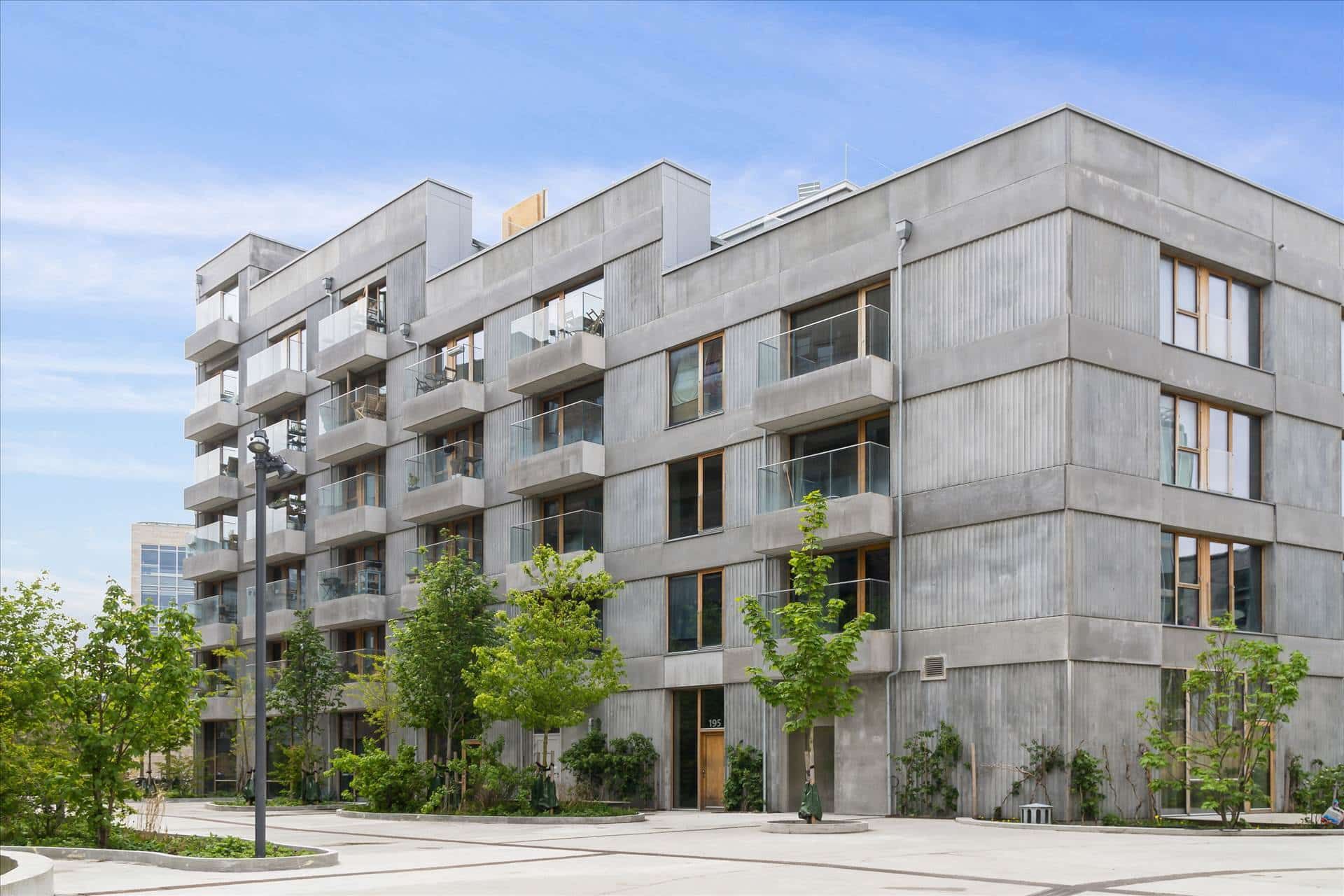 Lejlighed med store glasvinduer og terrasser, omgivet af grønne planter og træer, med en stor bygning og en travl gade i