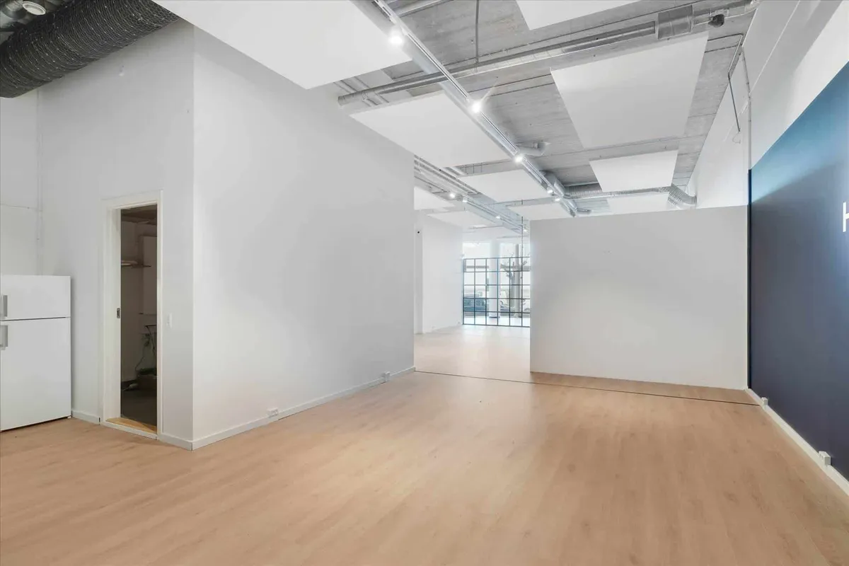 Rummelig og lys stue med hvidt loft, hvide vægge og trægulv. Døren er åben og viser en køleskab i et mindre rum.