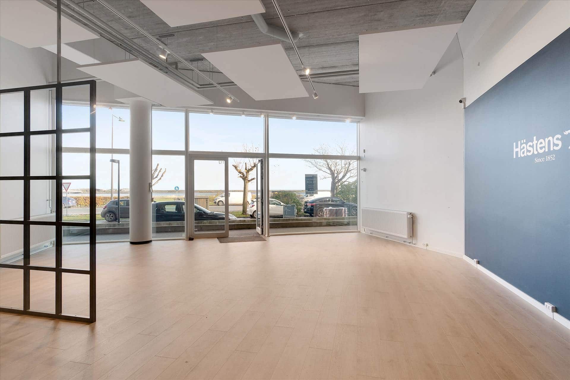 En stor indgangsdør med glas og metal, hvor der er et Hastens skilt på væggen og parkerede biler udenfor