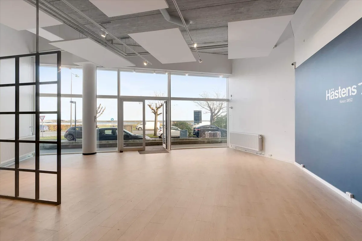 En stor indgangsdør med glas og metal, hvor der er et Hastens skilt på væggen og parkerede biler udenfor