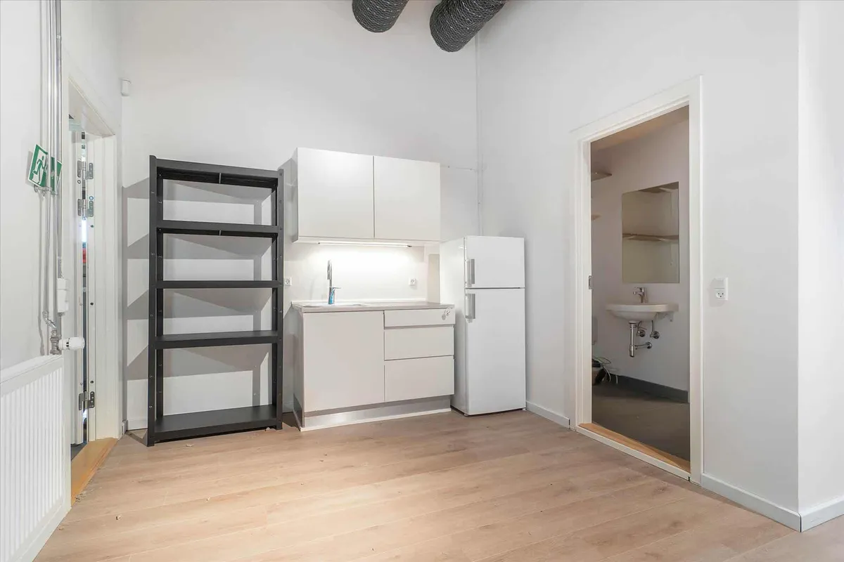 En køkken med hvide vægge, et højt hjørneskab og en hvid køleskab med håndtag samt en håndvask med spejl