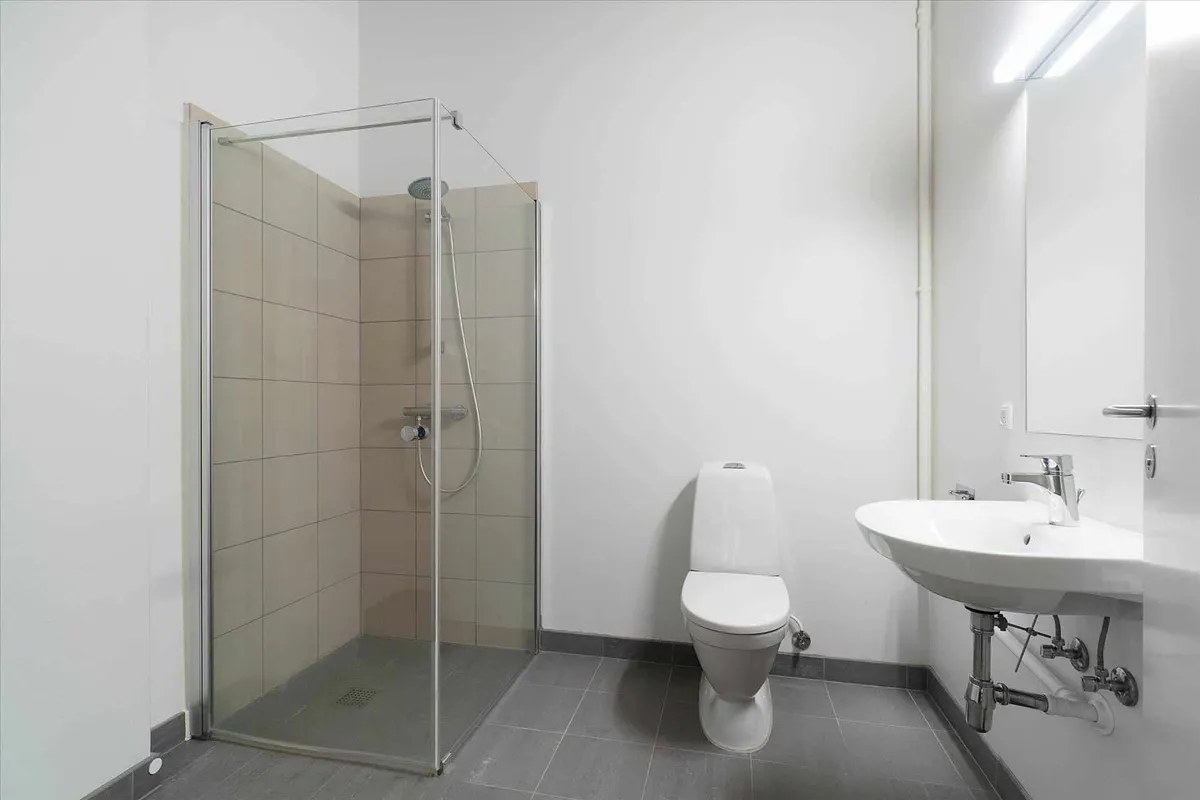 En lille, lyse badeværelse med et hjørnebruser og et toilet. Badestedet er udstyret med hvide kakel og en hvid håndvask med