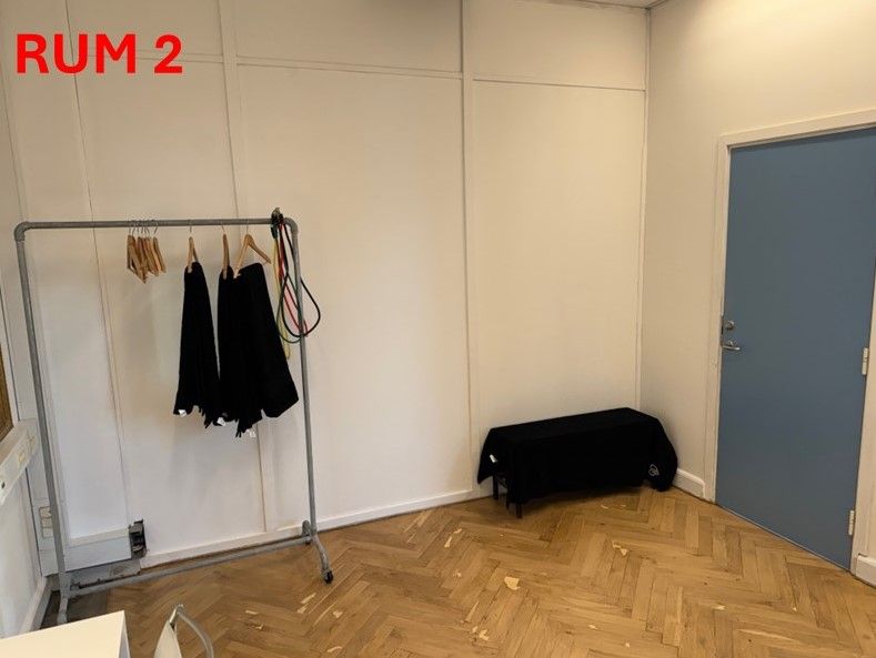 Et tomt rum med hvide vægge, trægulv og en hængekage med to sorte tøjstykker. Rummet har en lyseblå dør og en sort bord i