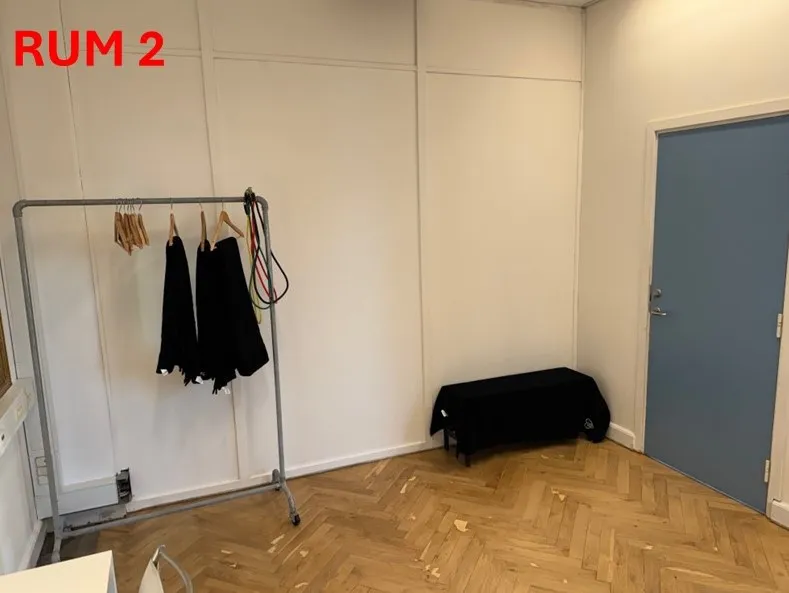 Et tomt rum med hvide vægge, trægulv og en hængekage med to sorte tøjstykker. Rummet har en lyseblå dør og en sort bord i