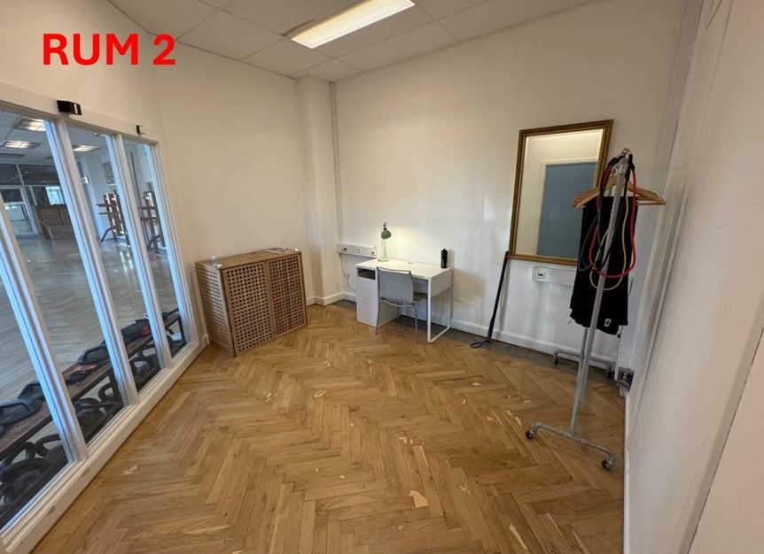 Foto af en tom rum med hårdt trægulv og hvide vægge og loft. Bord med lampe og stol. Spejl med tøjstativ med klæder.