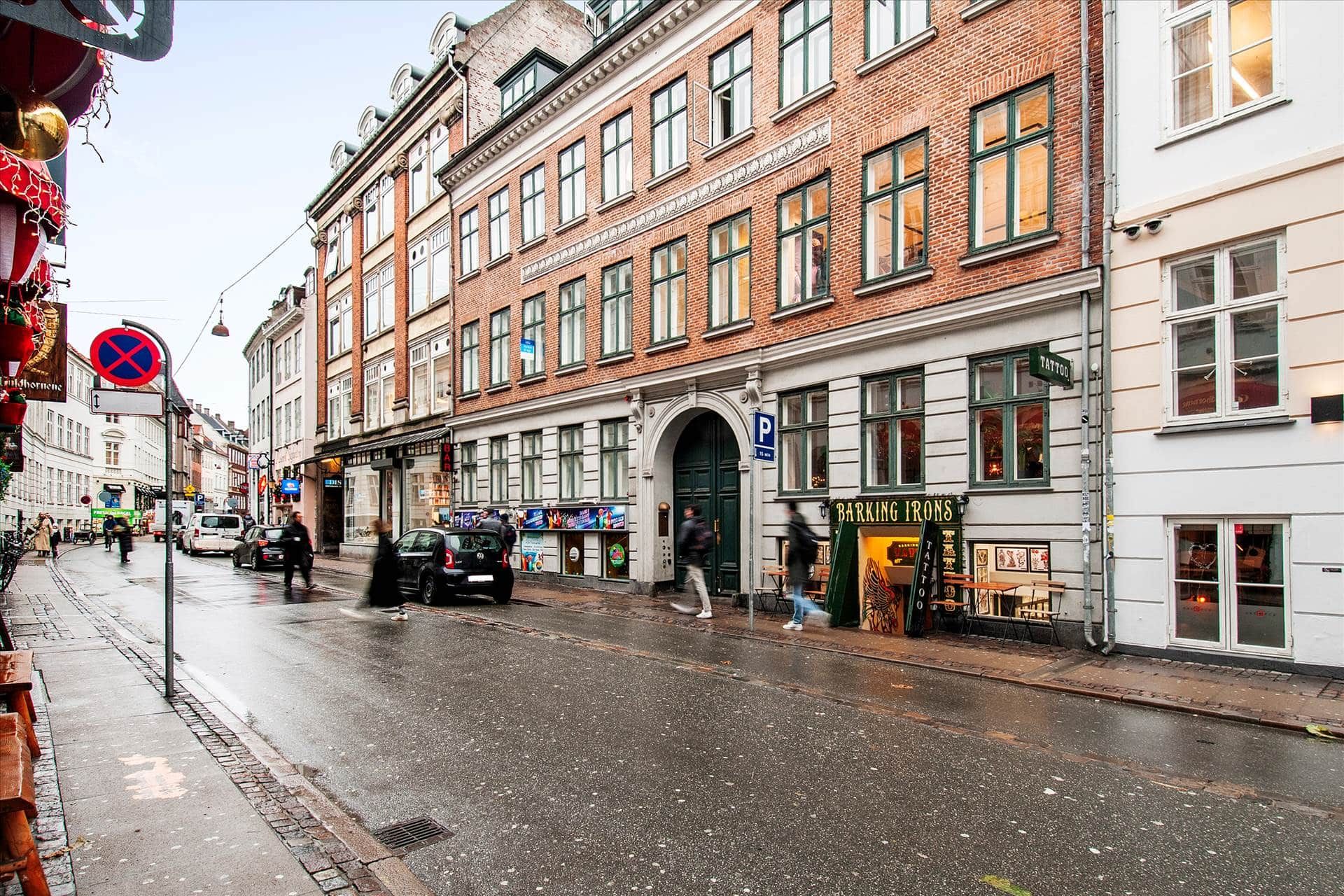 Vej med folk og biler, et rødt og blåt skilt, bygninger med mange vinduer og en vejledningsskilt for parkering.