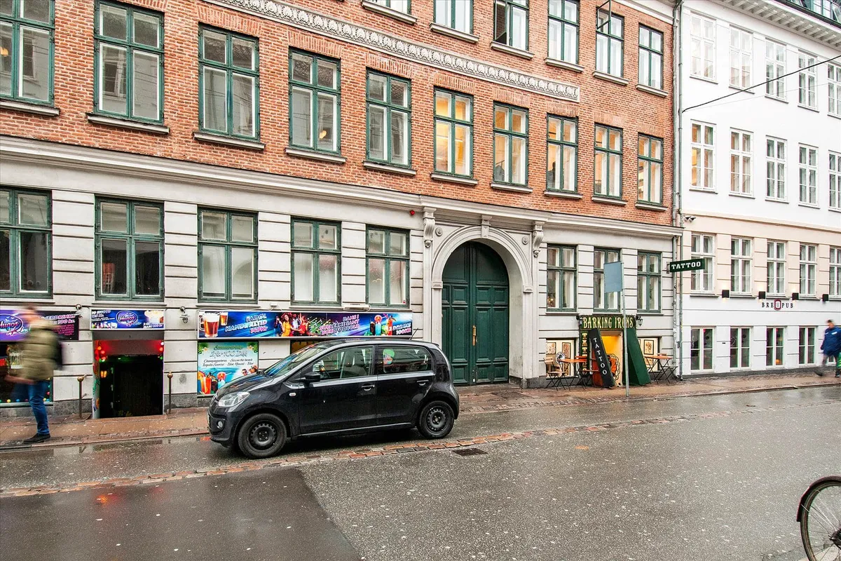 En smuk bygning med røde mursten og grønne ruder ligger på en regnfuld dag. En sølvmærket bil er parkeret på gaden. To