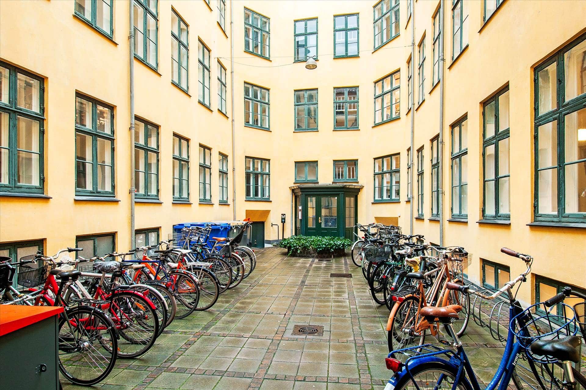 Række af cykler parkeret i gadeforhøjning med gule lejlighedsbygninger i baggrunden, og en grøn indgangsdør med søjler
