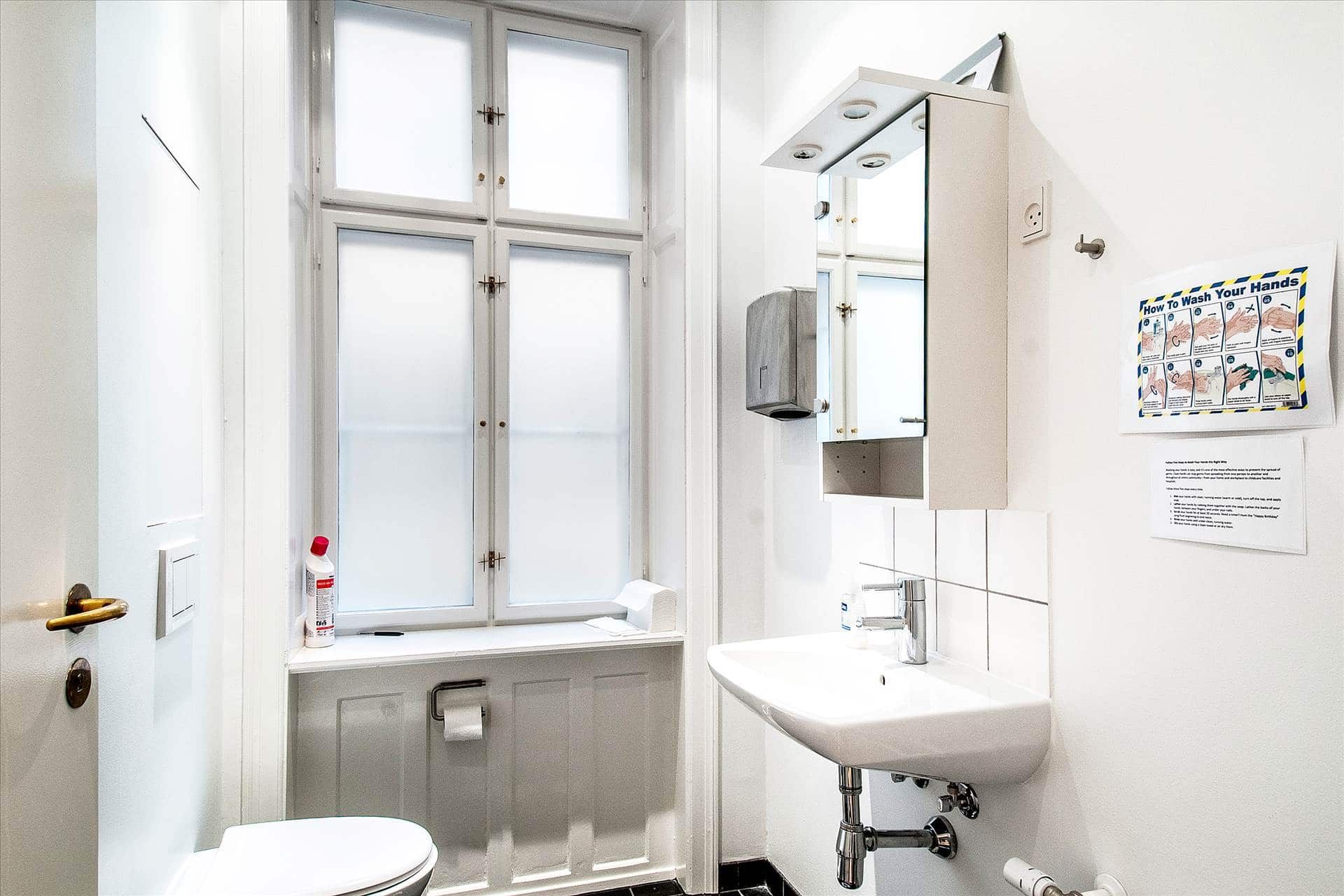 Hjørnebadeværelse med hvid toilet, hvidt skab med spejl og håndvask samt en håndvaskvejledning på væggen.