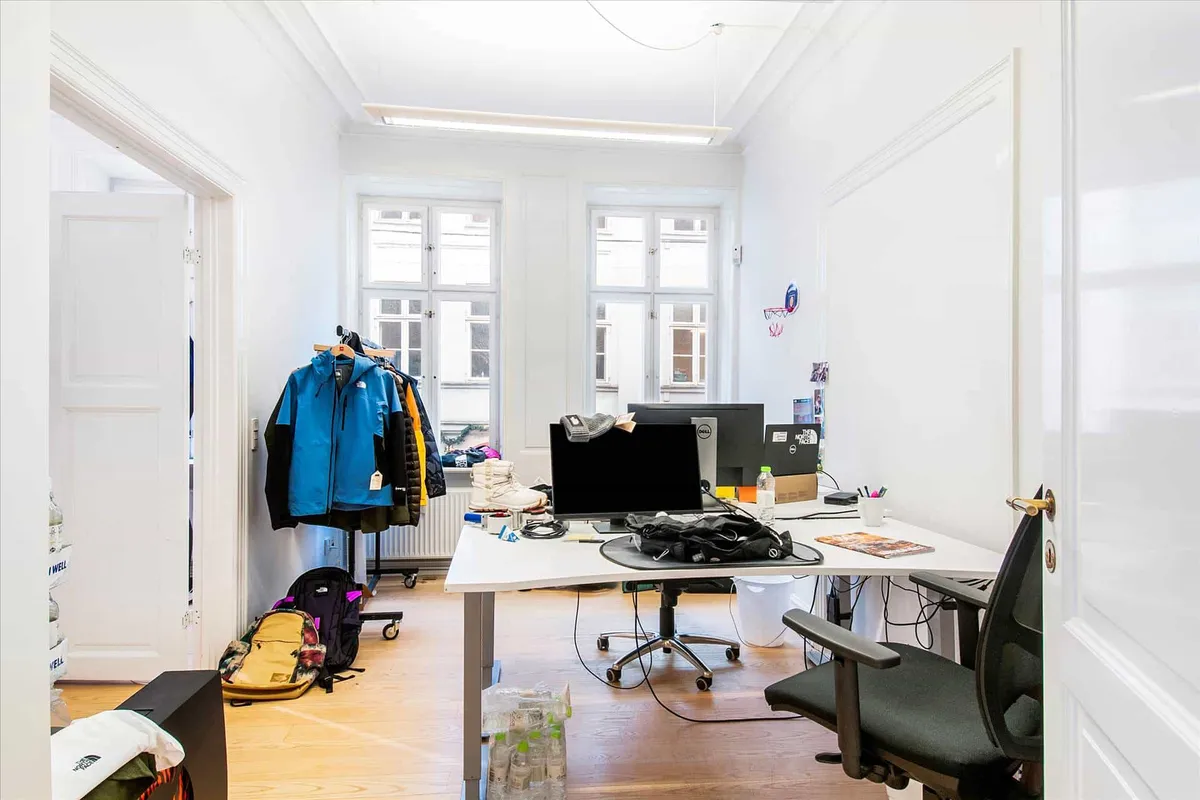 Lejlighed med to skriveborde, skrivebordsstole og computere samt to jakker hængende på en klædeskab.