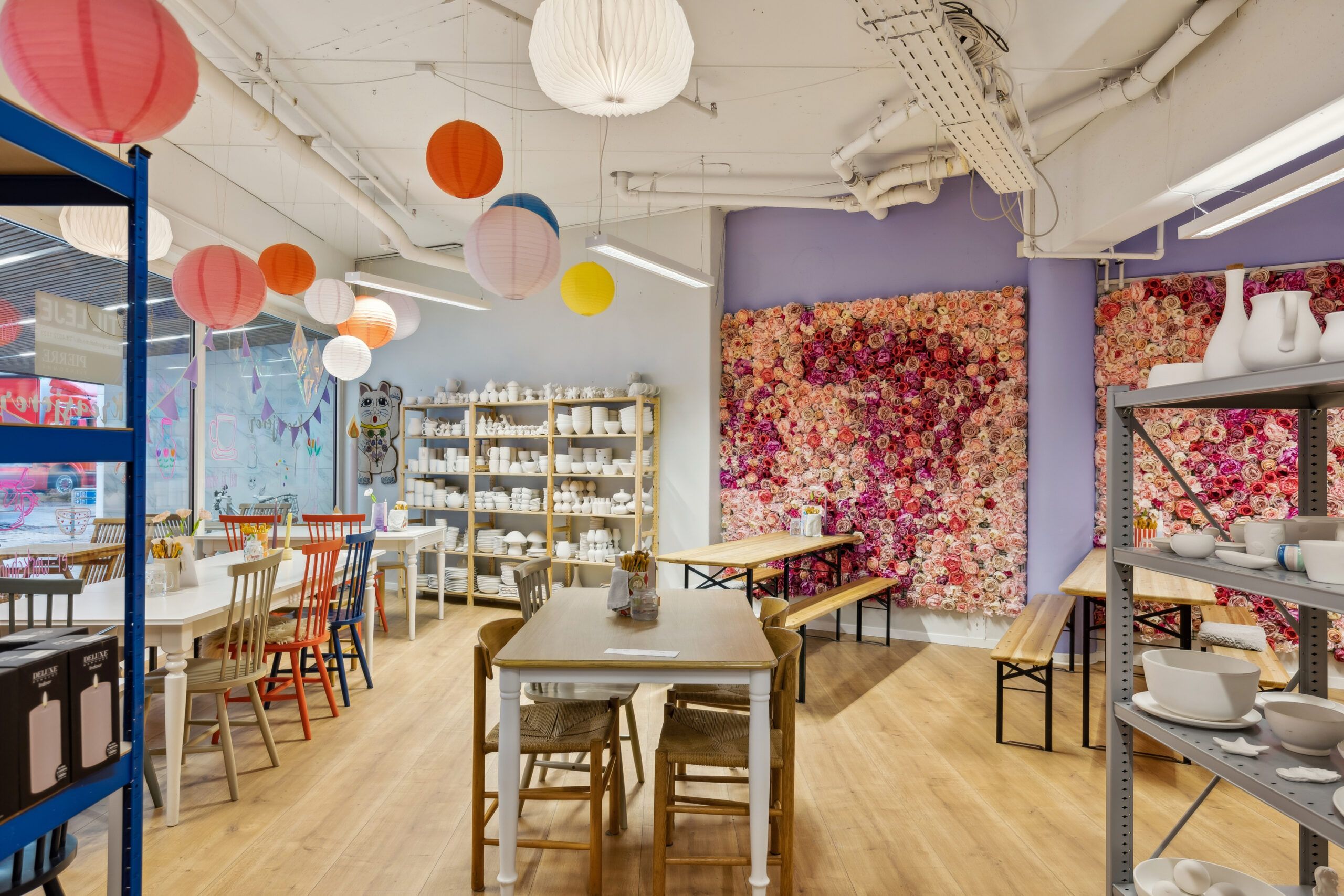 Inde i en farverig og moderne indretning med lyse farver, lysekroner, og blomstervæggen. Bord og stole står foran en hylde