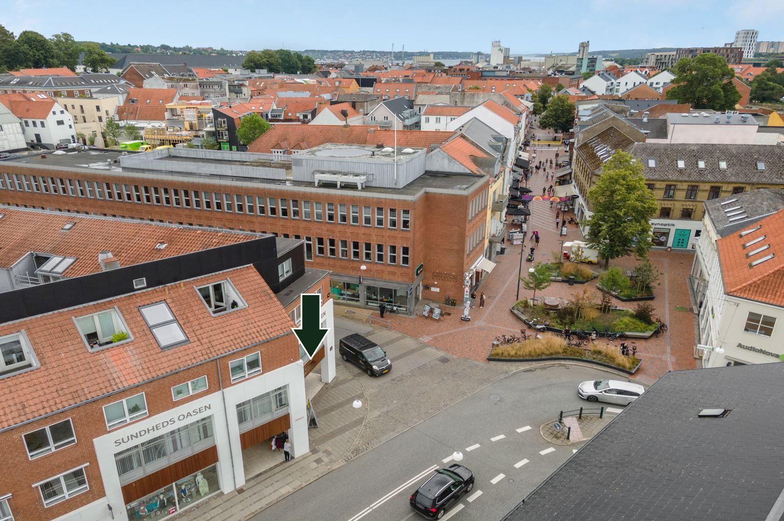 Byudsigt med mange huse og gader, flere biler og mennesker, hjørnebygning med skilt SUNDHEDS OASEN