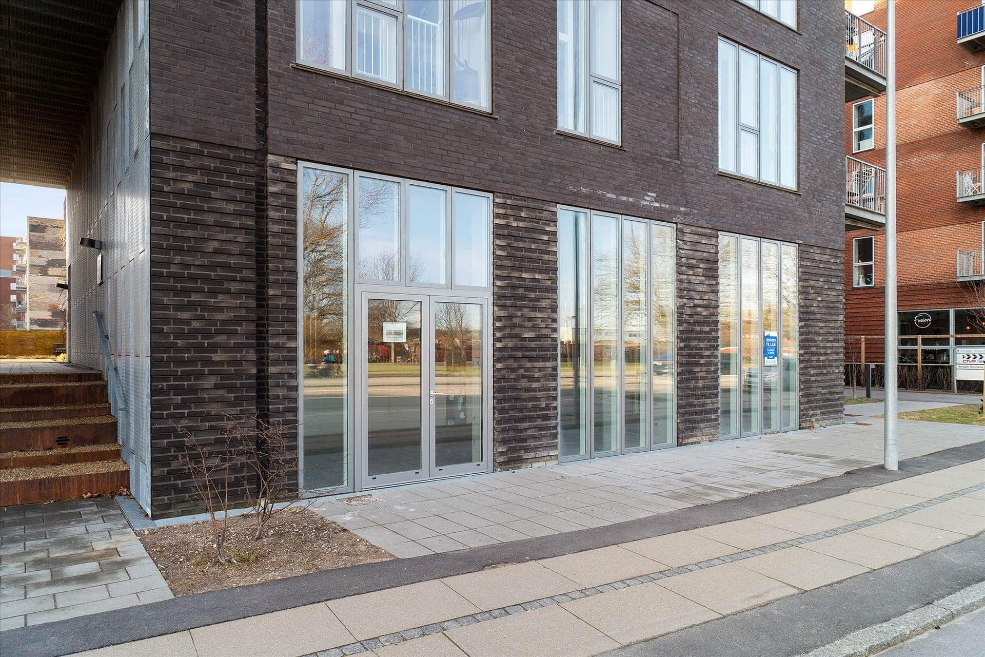 Moderne erhvervslokaler med glasdøre og vinduer og et terrasseområde med skurtag, der vender ud mod en gågade med