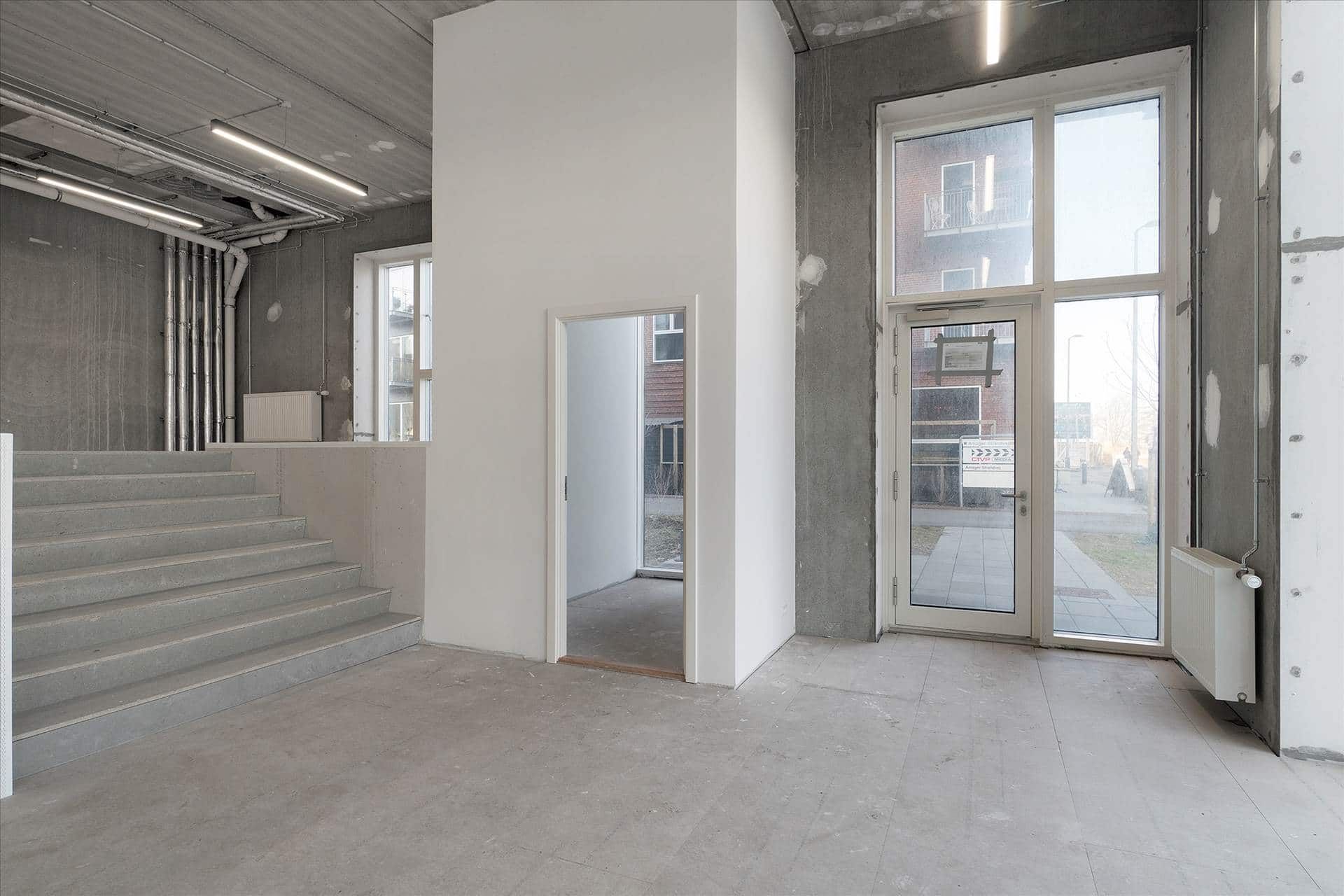 Lokaler med højt til loftet og store vinduer. En hvid væg og en dør med glas i samme farve. En forside med lysegrå