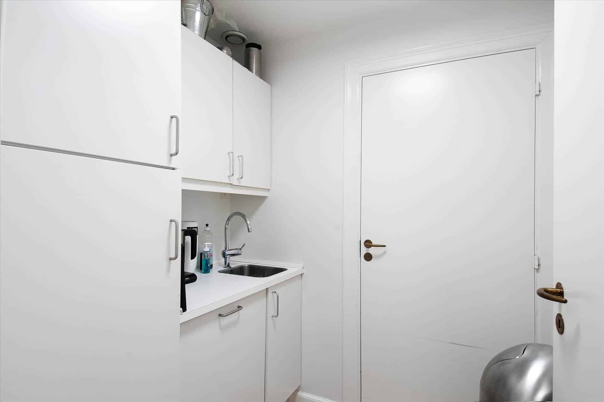 En lille køkken med hvide vægge og hvide skabe, en håndvask og et køleskab med en lyseblå flaske og en sprayflaske på den