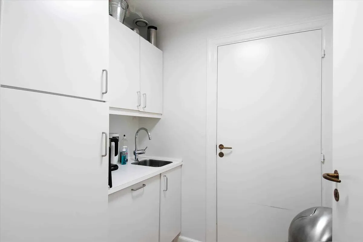 En lille køkken med hvide vægge og hvide skabe, en håndvask og et køleskab med en lyseblå flaske og en sprayflaske på den