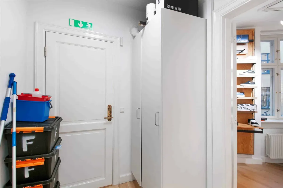 Lagerrum med sort og blå opbevaringsboks og rengøringsudstyr samt et hvidt skab og en åben dør til et område med skoskabe