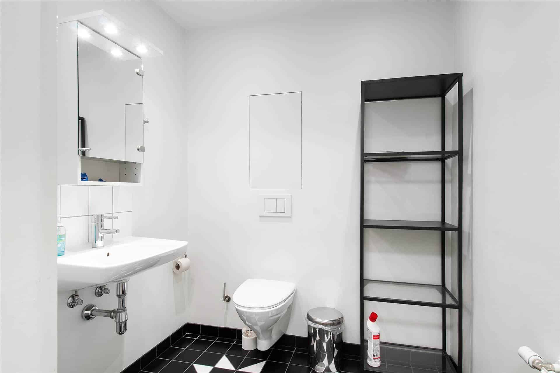 Minimalistisk badeværelse med hvidt toilet, hvidt vask og sort-hvid gulvfliser. Der er et højt sort hyldesystem og en hvid