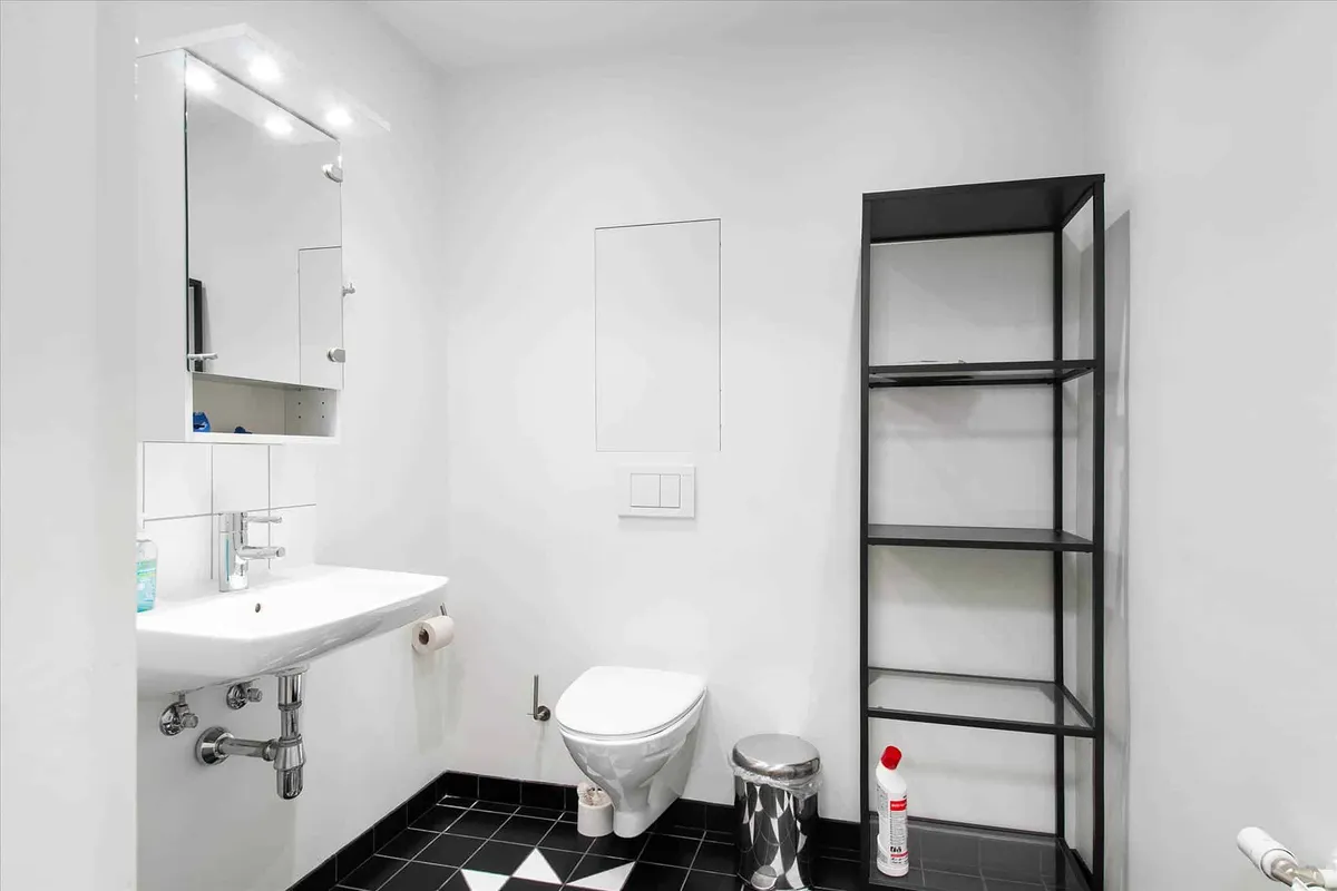 Minimalistisk badeværelse med hvidt toilet, hvidt vask og sort-hvid gulvfliser. Der er et højt sort hyldesystem og en hvid