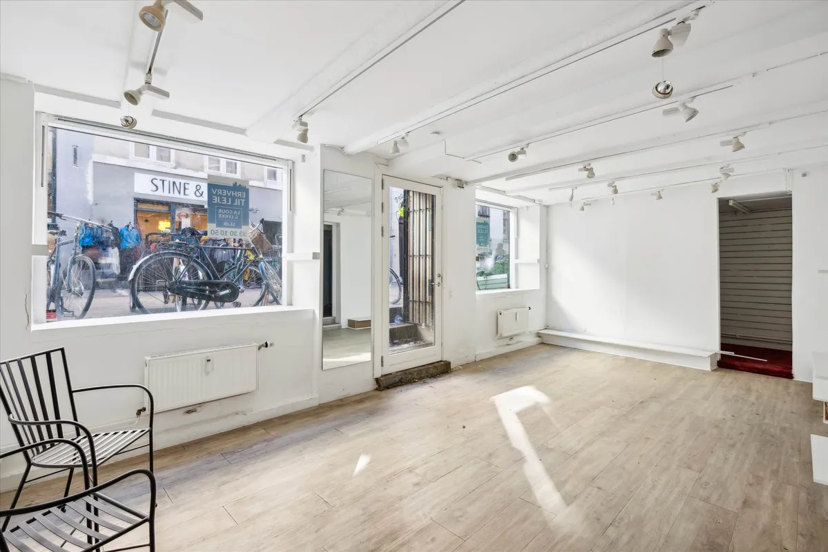 Et tomt rum med hvide vægge, lysekroner og trægulve. Et spejl og en dør med glas og metal. En stol og en radiator er