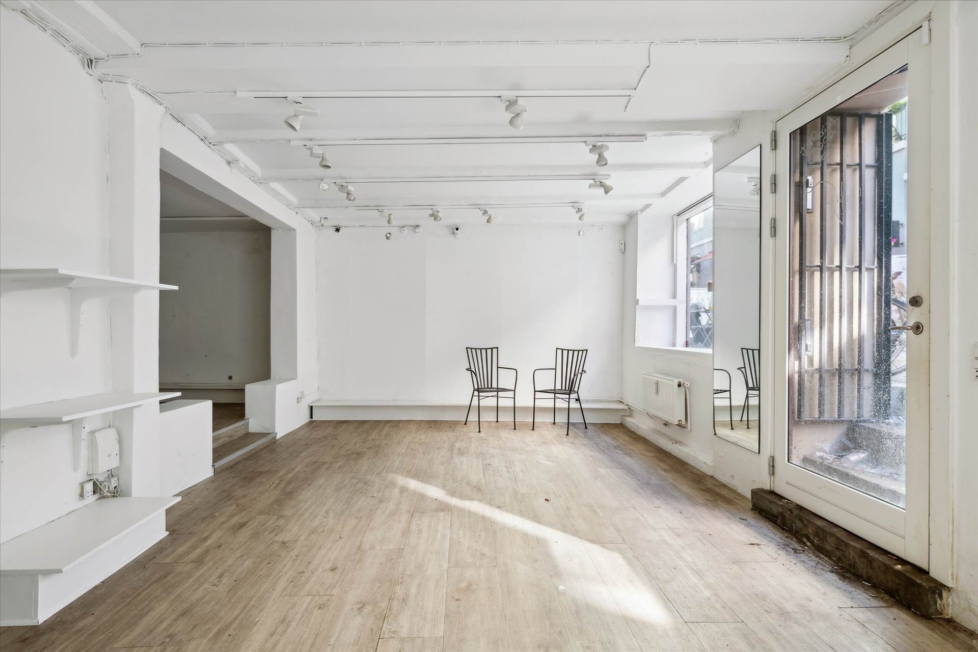 To stolpe med hængslede døre og spejle samt to stole i en hvid rummeligt lokale med lysegul trægulv og hvide vægge.