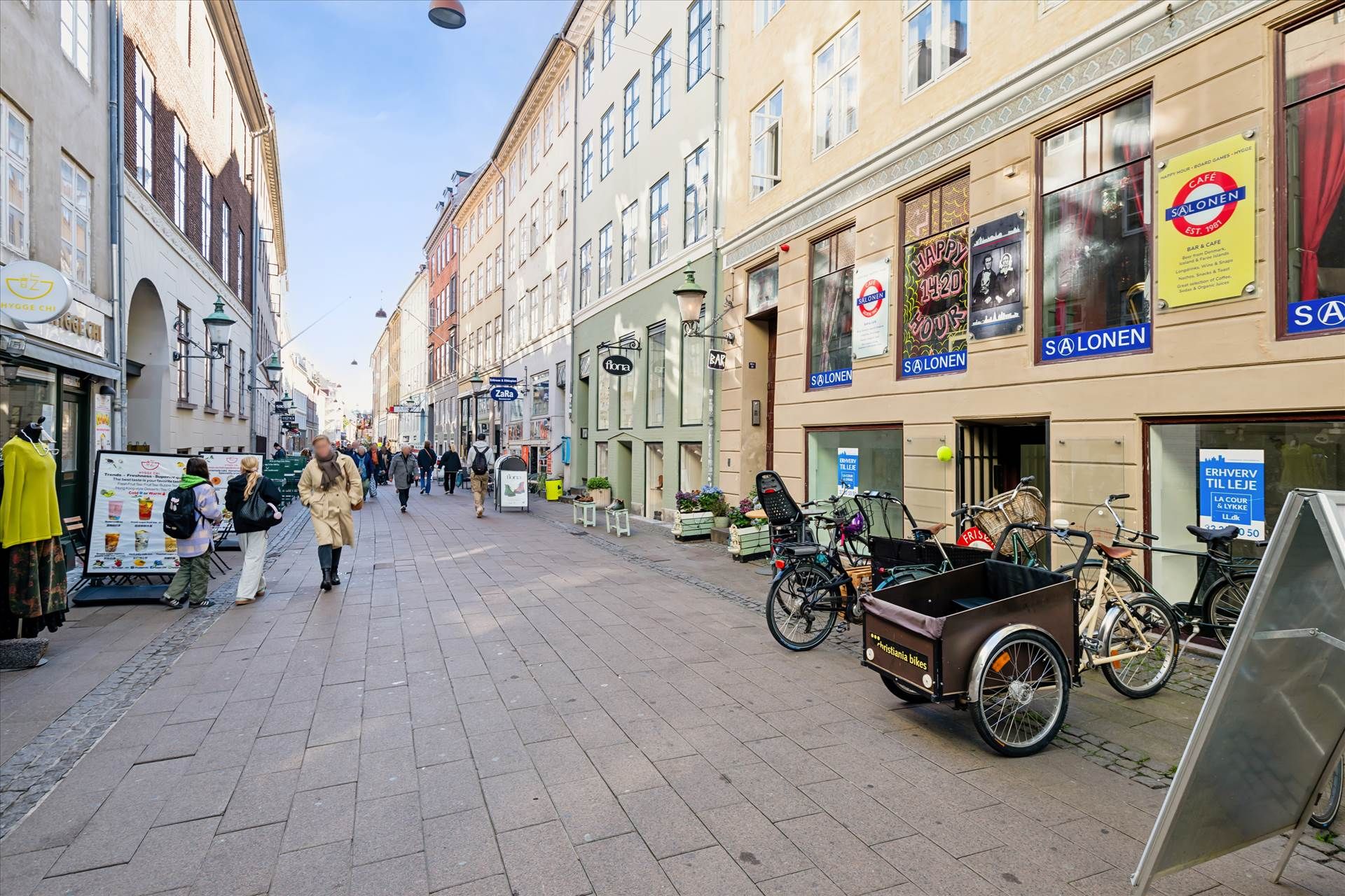 Gade med gågade, hvor folk går og ser på butikker. Butikker med udsalg og reklamer. Cykler og lastcykel står udenfor