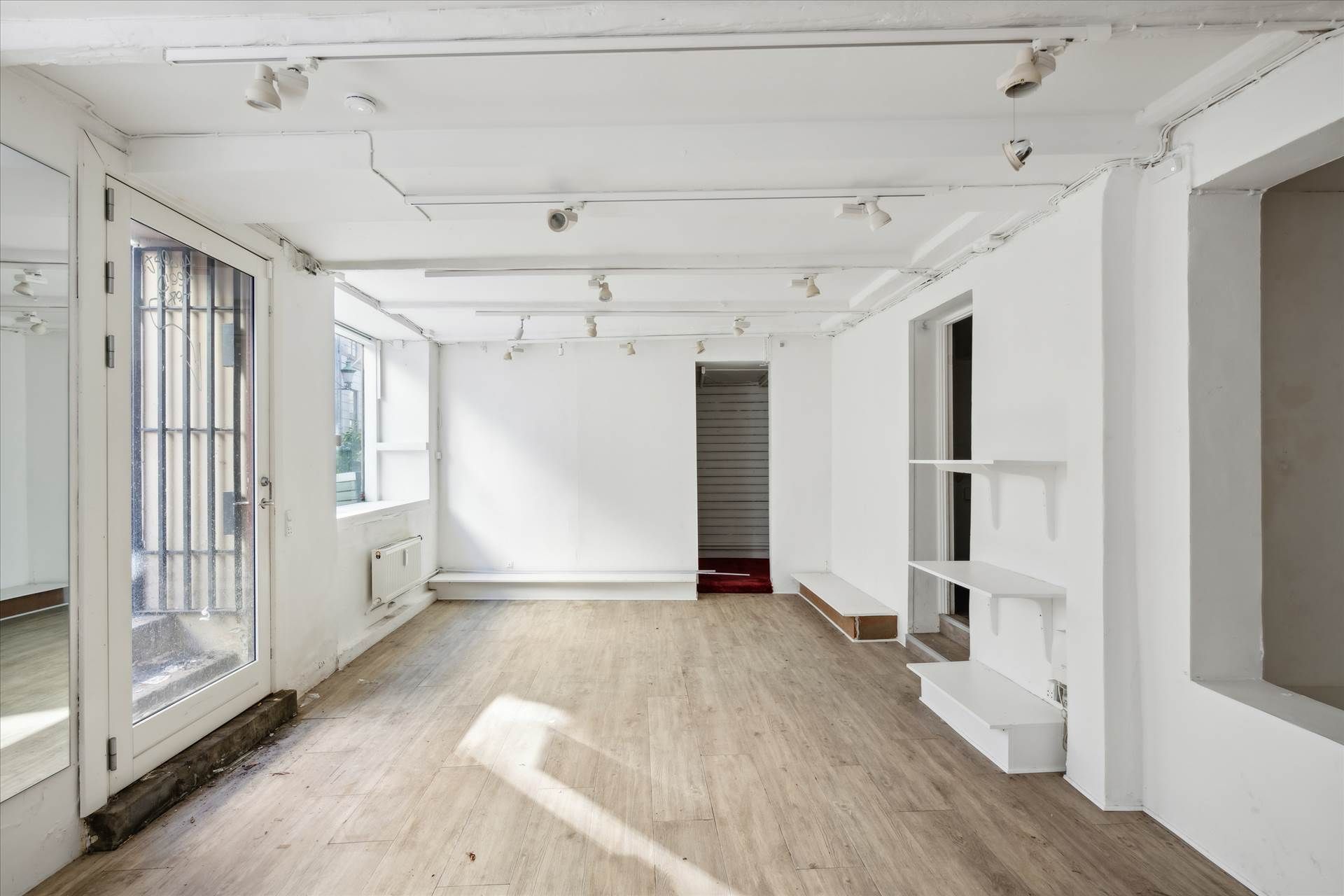 Et tomt rum med lysegult gulv, hvide vægge og slanke hjørnehylder. Der er to vinduer og en dør med metalruder og håndtag