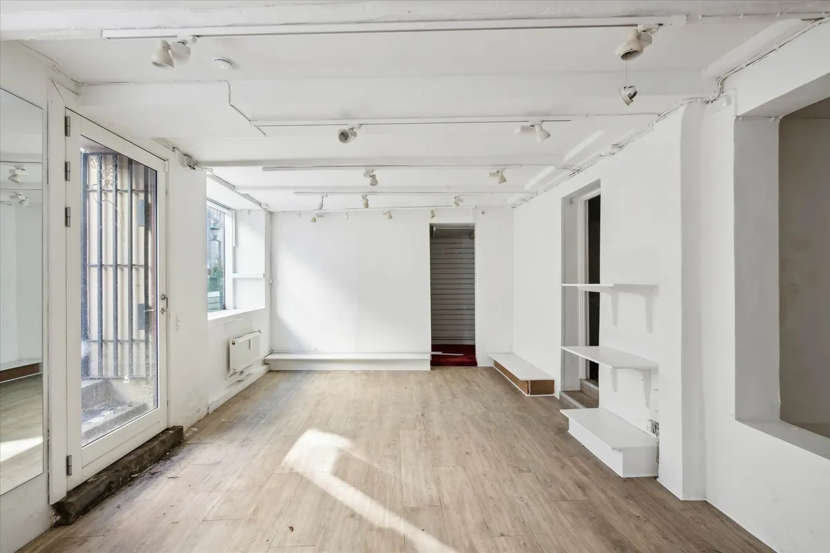 Et tomt rum med lysegult gulv, hvide vægge og slanke hjørnehylder. Der er to vinduer og en dør med metalruder og håndtag