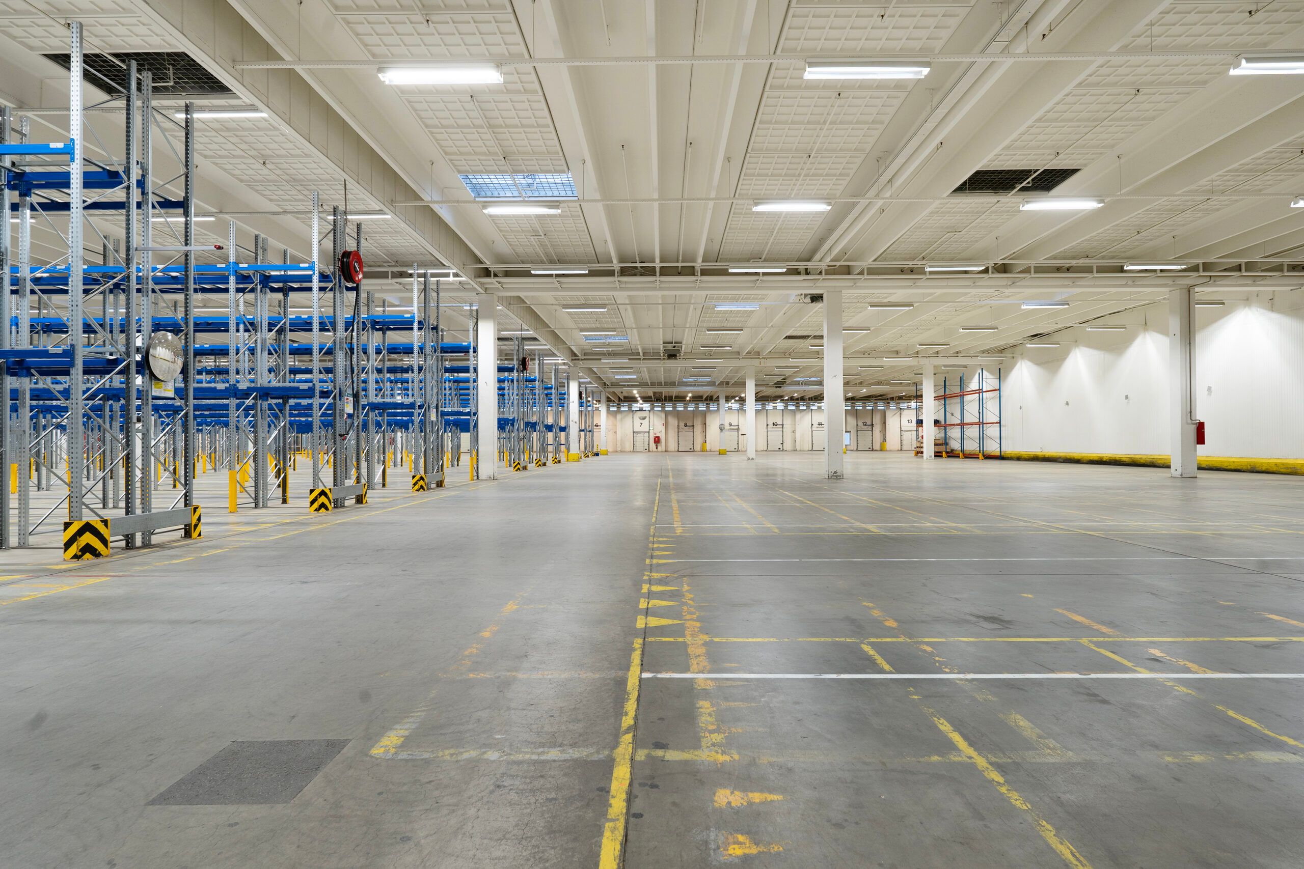 Lager med flere rækker af paletter. Gulv og loft er hvide, og der er markeringer på gulvet og gule streger på loftet.