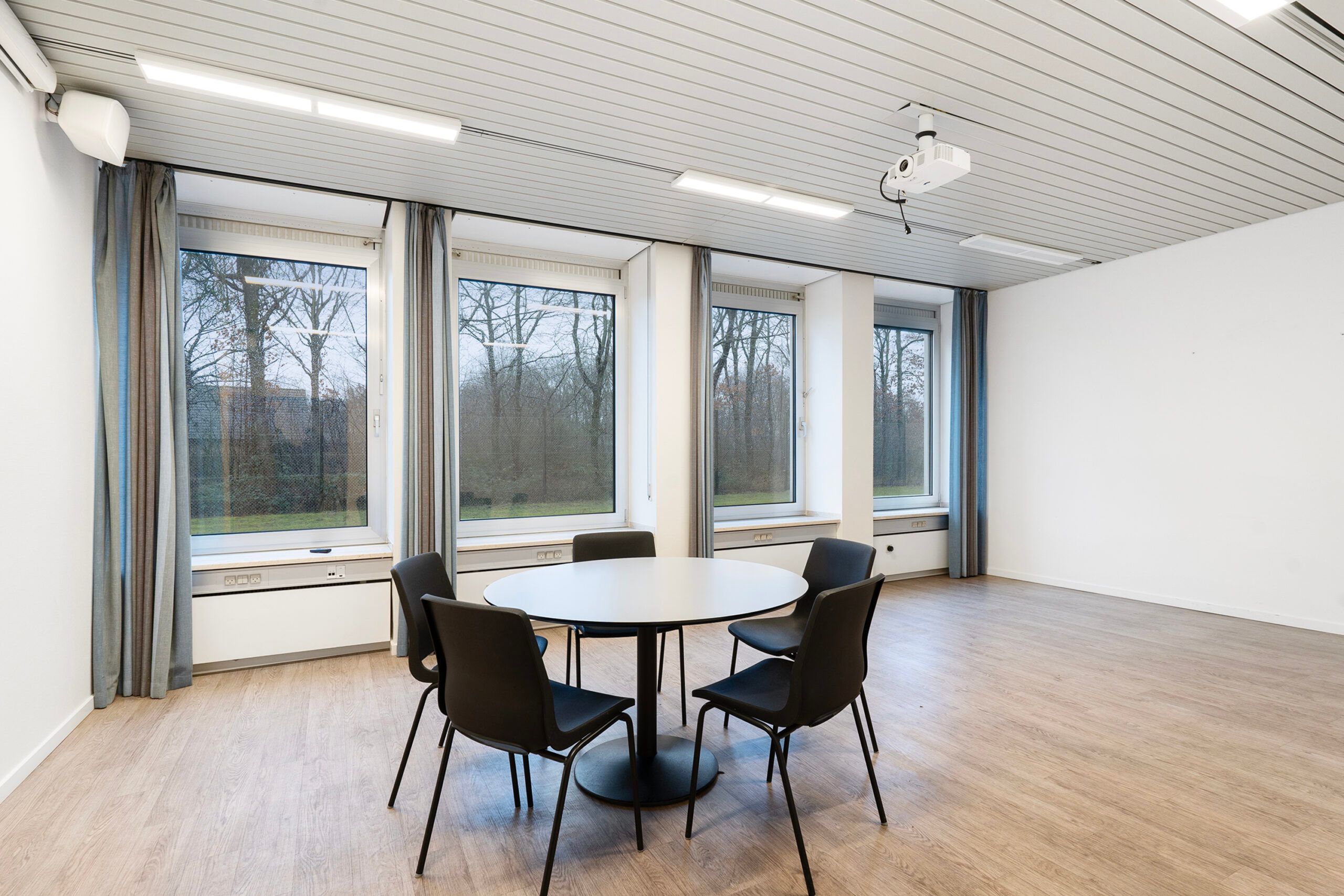 Rundt bord med fem stole i mødelokale med hvide vægge, højt gulv og vinduer med persienner og udsyn til skov