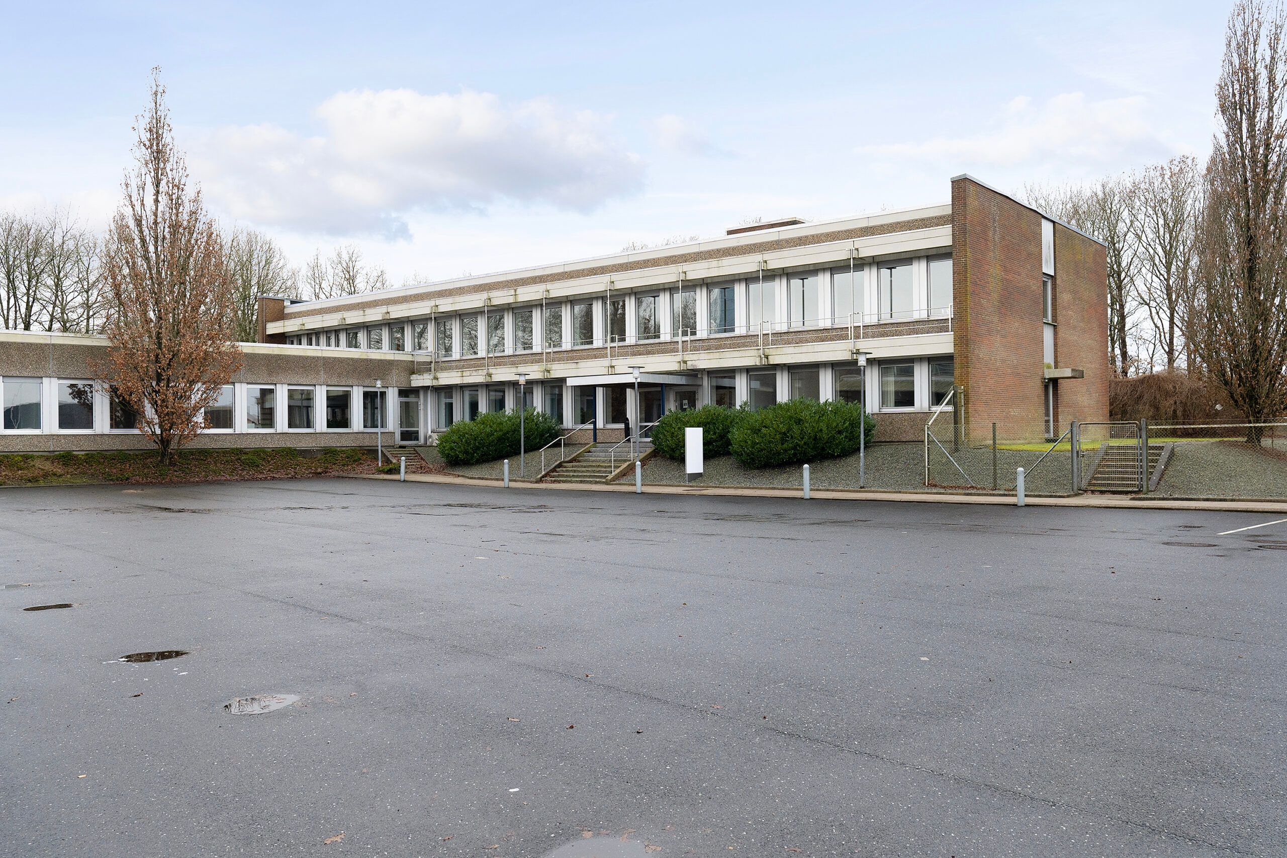 Et stort bygningskompleks med glasruder og terrasser og eksteriør i brun mursten. Større område foran med skrapeløse veje og