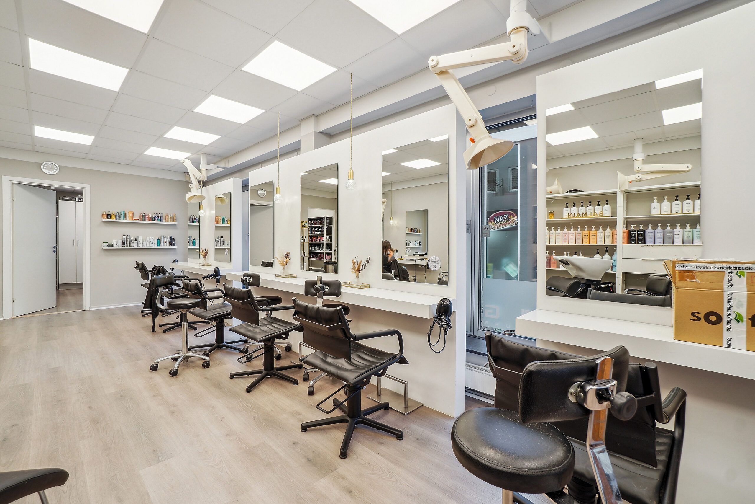 En ren og moderne frisørsalon med højloftede og hvide vægge, lyse loftsbelysning og lysebrune gulvbelægninger. I salonen er