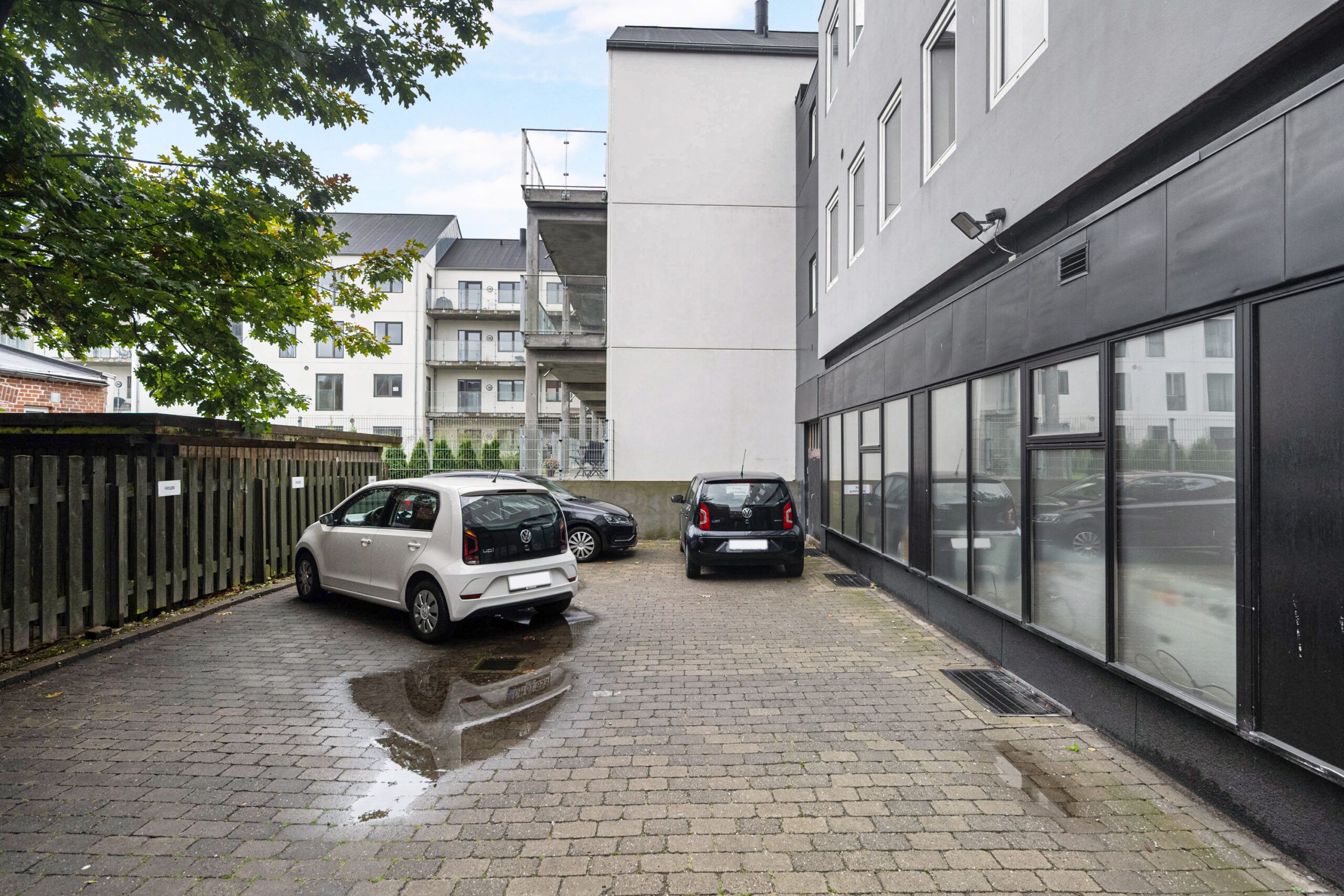 På en betonbelægget gade parkeret to biler og en tredje bil parkeret foran et bygningskompleks med glasruder og en