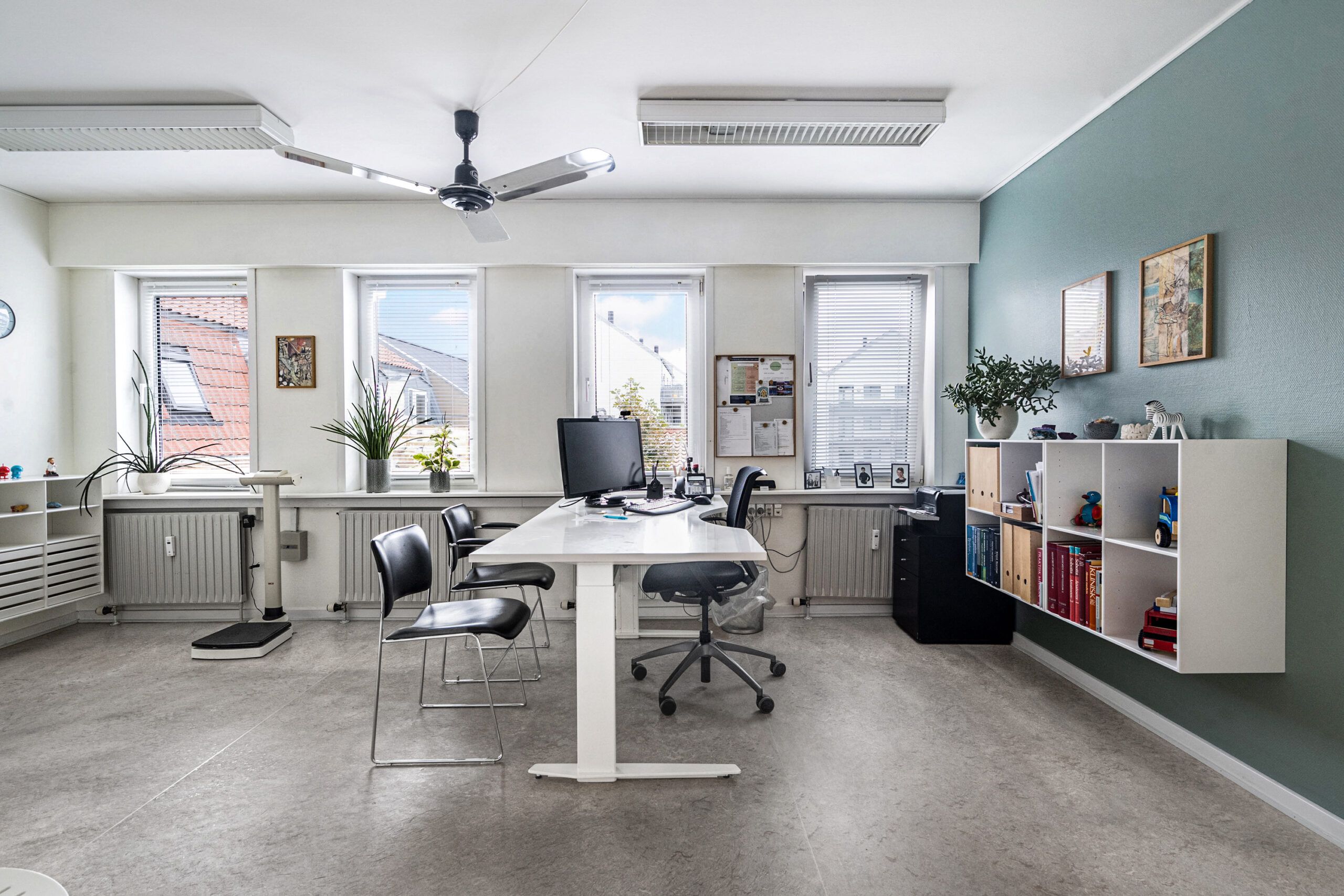 Kontor med højt loft, vinduer, tre skriveborde og to stol. Loftventilator og klimaanlæg samt plante i hjørne.