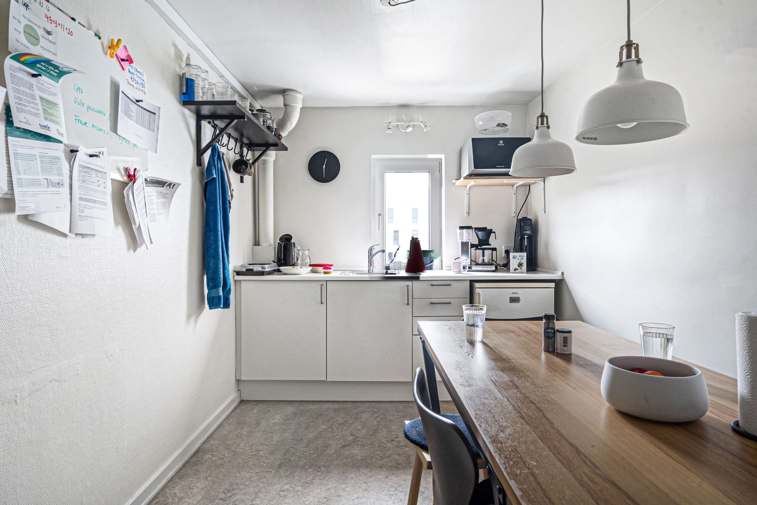 Køkken med hvidt køkken med skrald, to lysekroner, mikrobølgeovn, kaffemaskine, skrivebord, stole og glas med vand og frugt