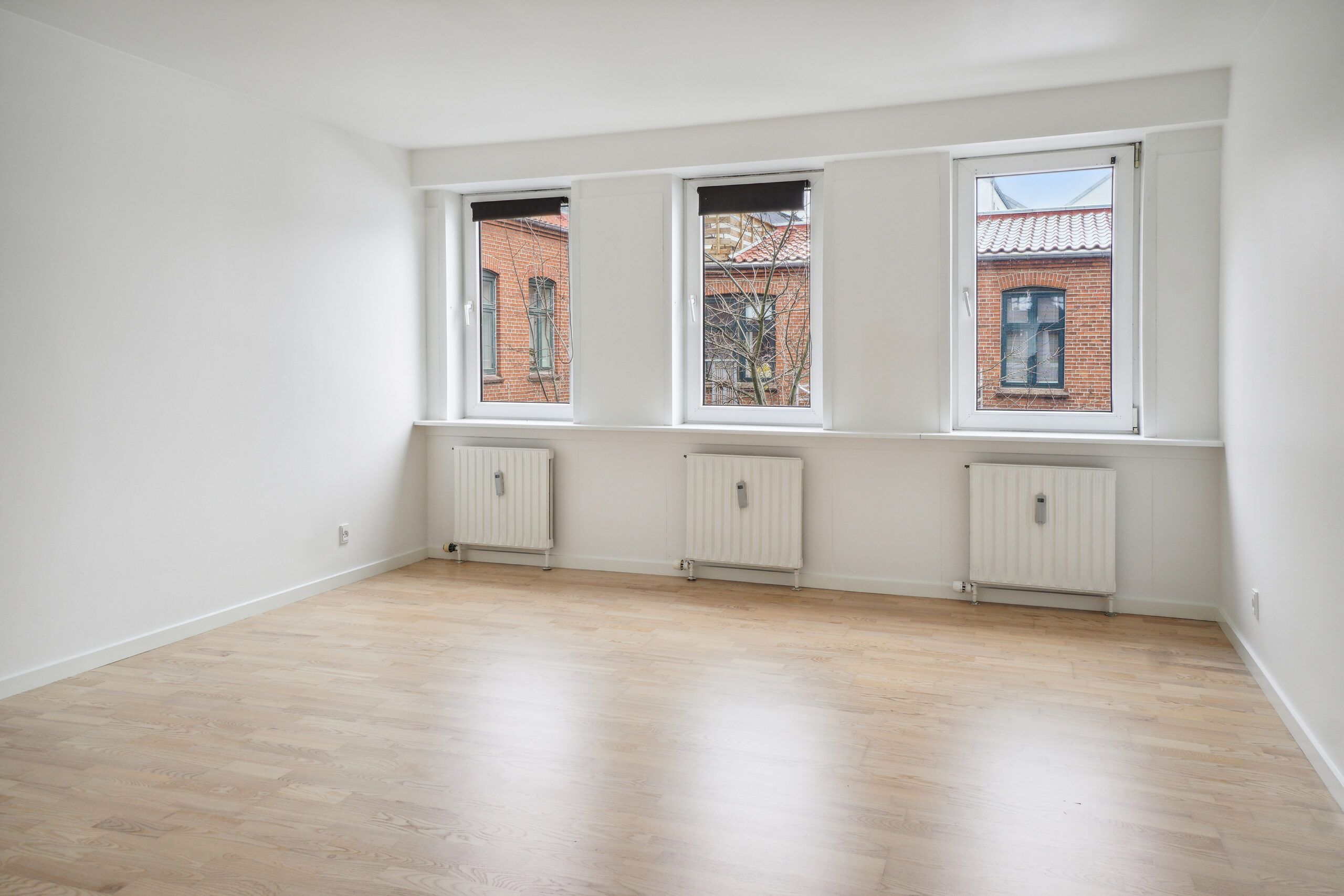 En tom lejlighed med hvide vægge og tre store vinduer med hvide ruder og jalousier