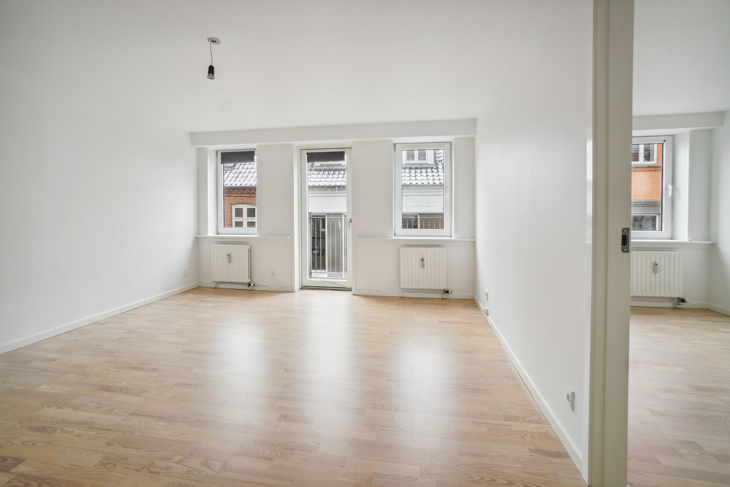 En tom lejlighed med hvide vægge og gulv, tre vinduer, radiatorer og et lysekroner hængende fra loftet.
