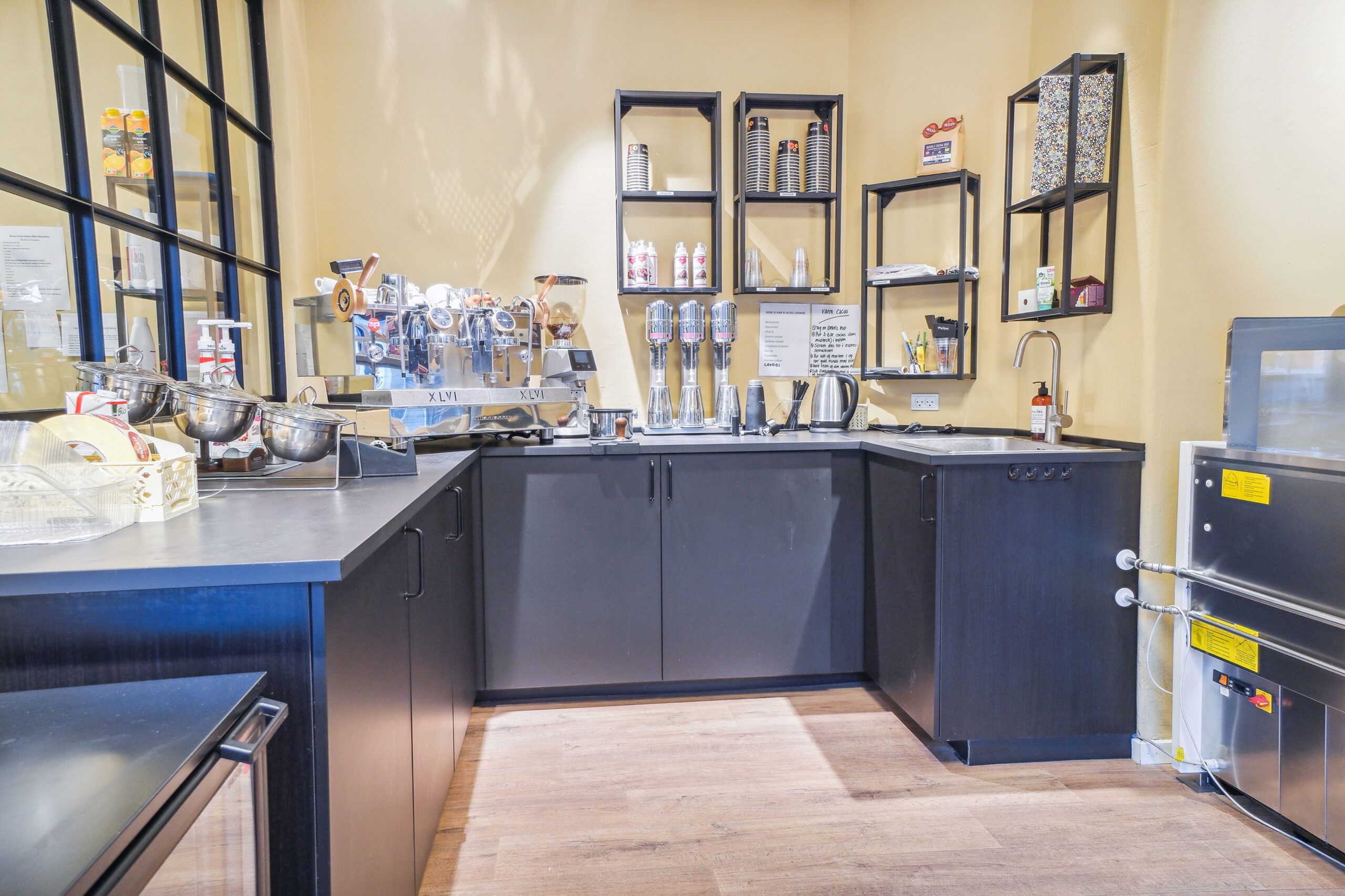 En køkkendeling med en række kaffemaskiner og flere apparater på hylder og bord. Der er også en håndvask med vandhane og en