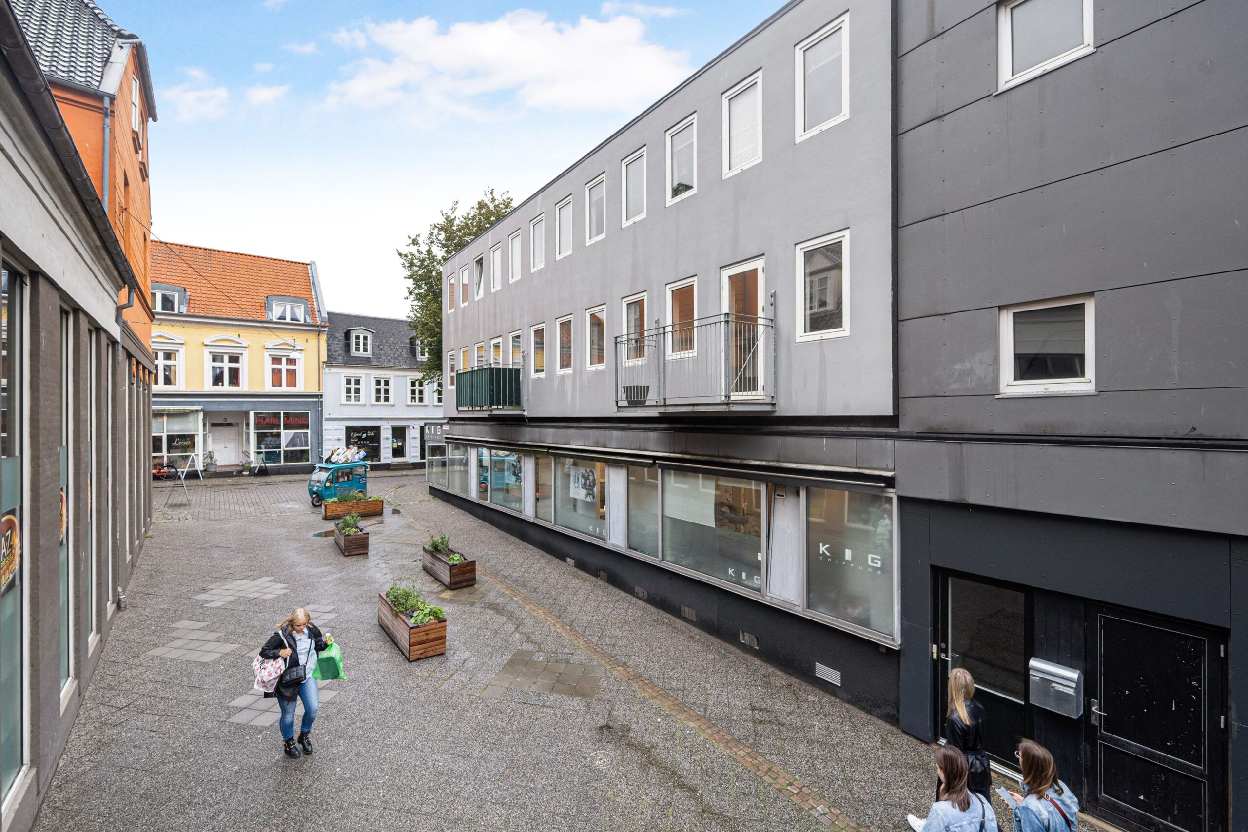 En kvinde med indkøbsposer går på en brolagt gade foran et stort bygningskompleks med butikker og lejligheder