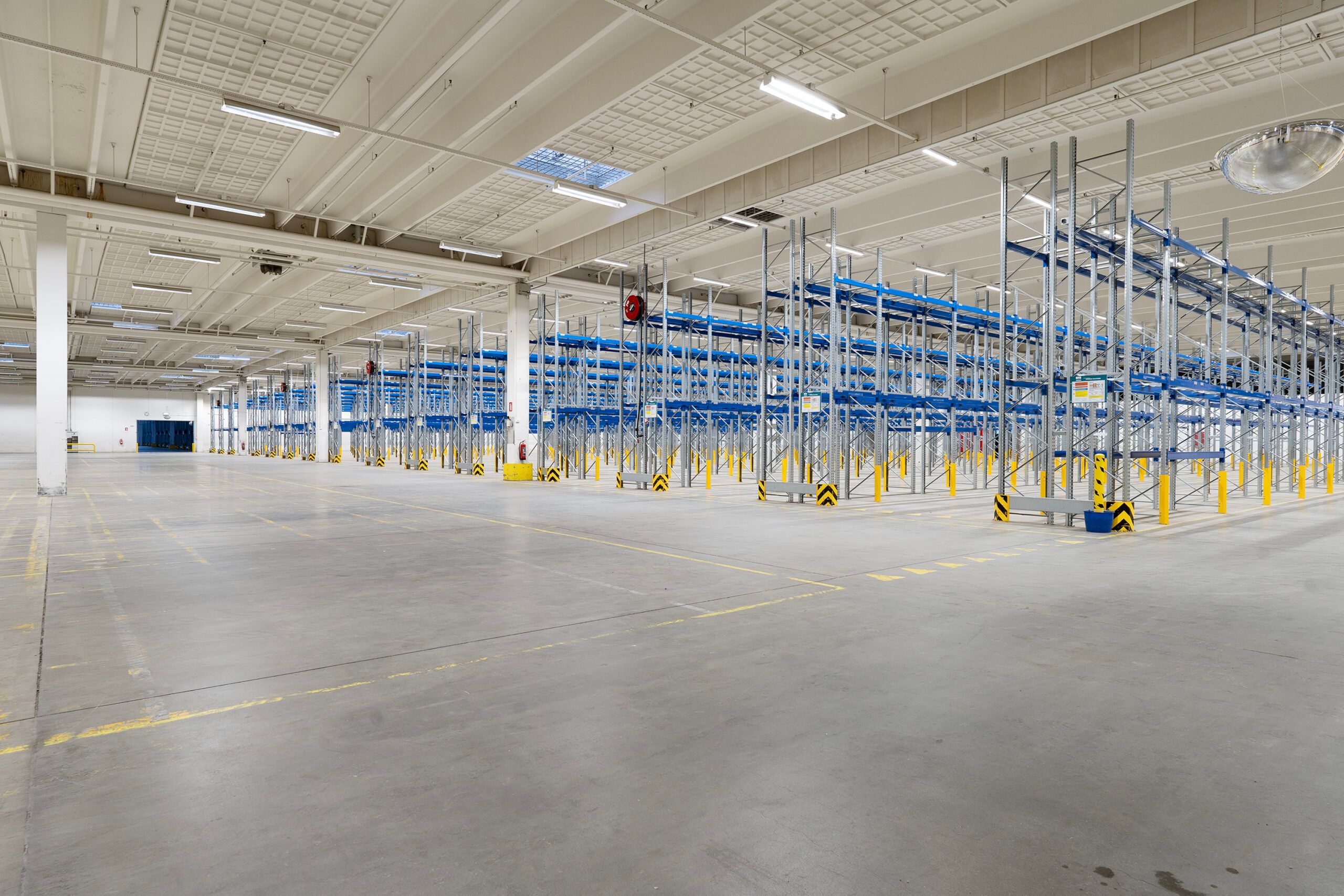Lager med flere rækker af blå lagerhylder og forskellige gulvmarkeringer og et rødt alarmknapper.
