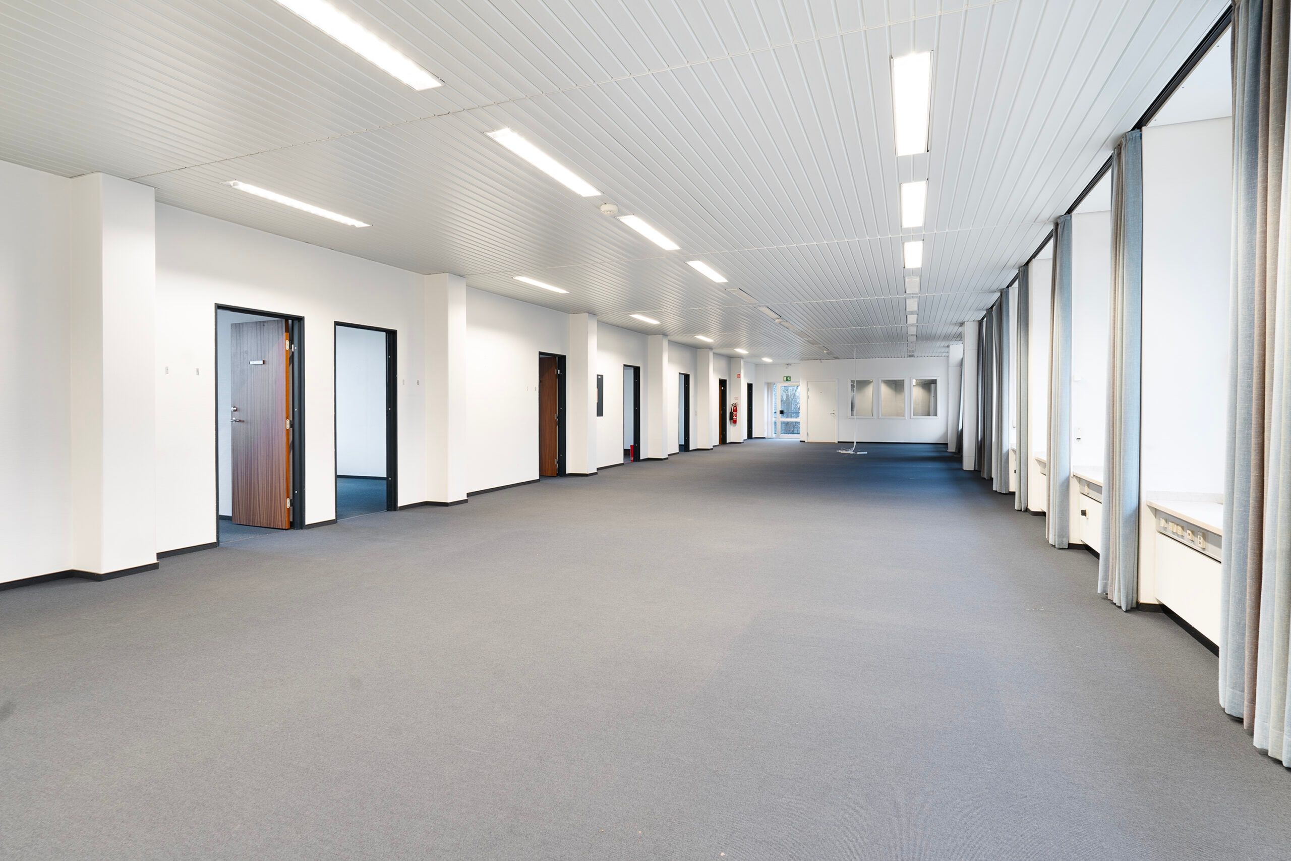 En tom, lyse hall med lysegult gulv og hvide vægge. Hallen er forsynet med flere døre og vinduer, samt lys på loftet.