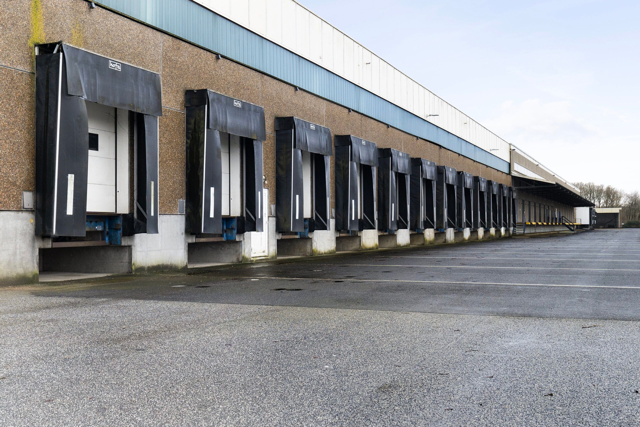 Lagerbygning med høje sorte skydeporte og hvide døråbninger og en stor, tom parkeringsplads med tydelige parkeringslinjer
