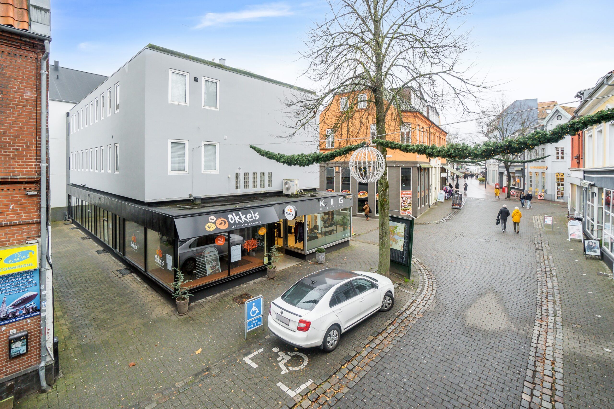 En hvid bil parkeret på en gågade i Kolding, med en skilt med handicapparkering, et hus med mange vinduer og et mærke for