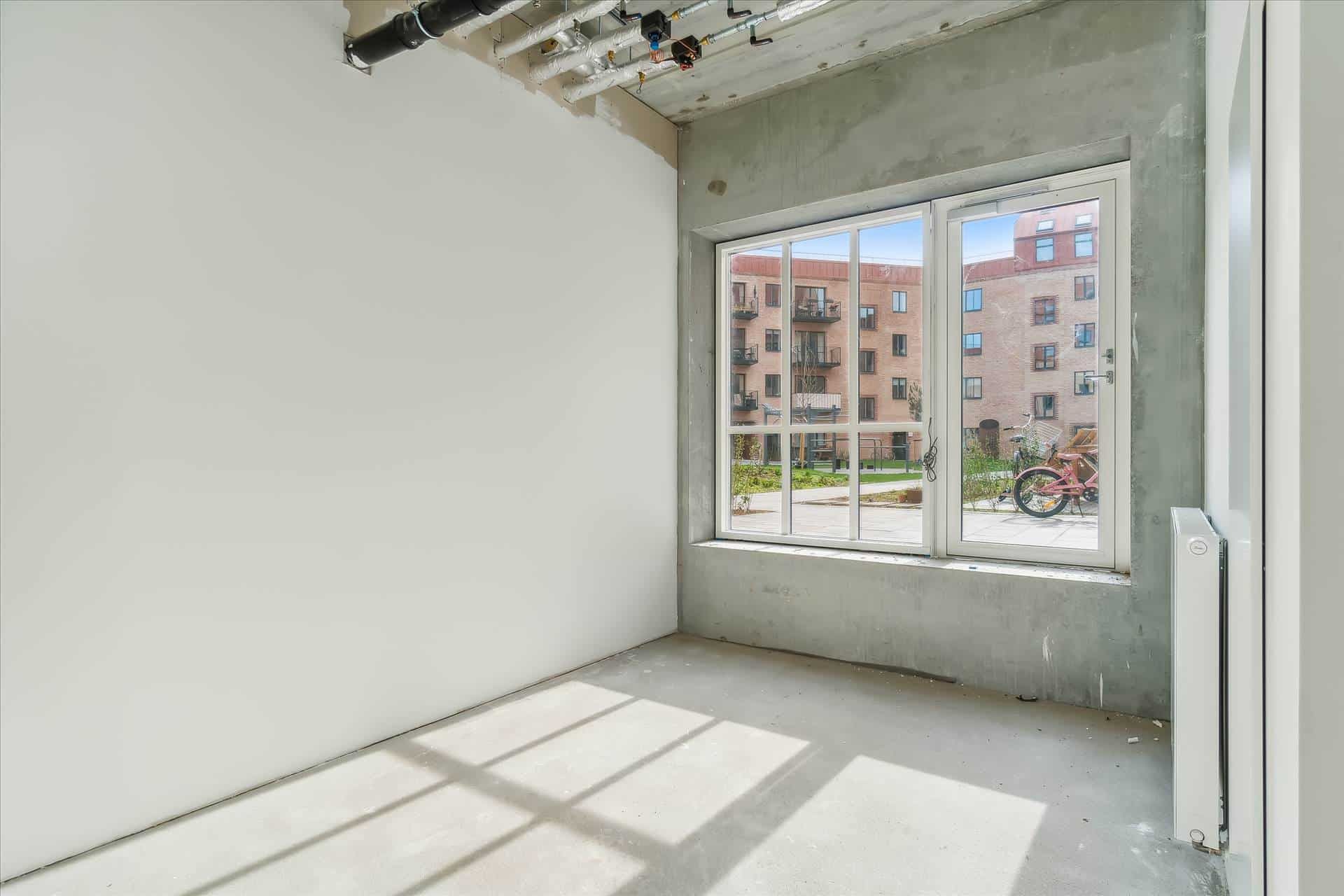 Et hjørneværelse med hvidt vægmaleri og glasvinduer med udsigt til park med bænke, træer og pink cykel i udkanten af bygning