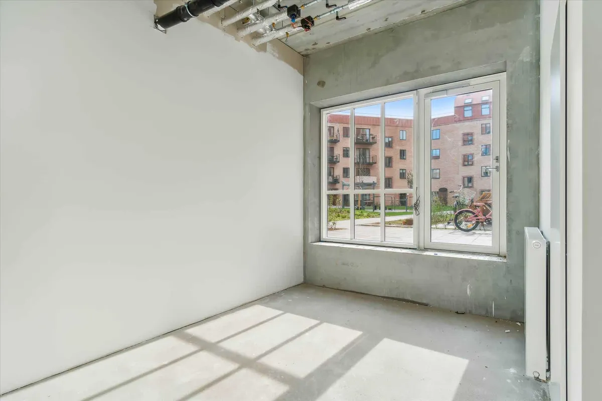 Et hjørneværelse med hvidt vægmaleri og glasvinduer med udsigt til park med bænke, træer og pink cykel i udkanten af bygning