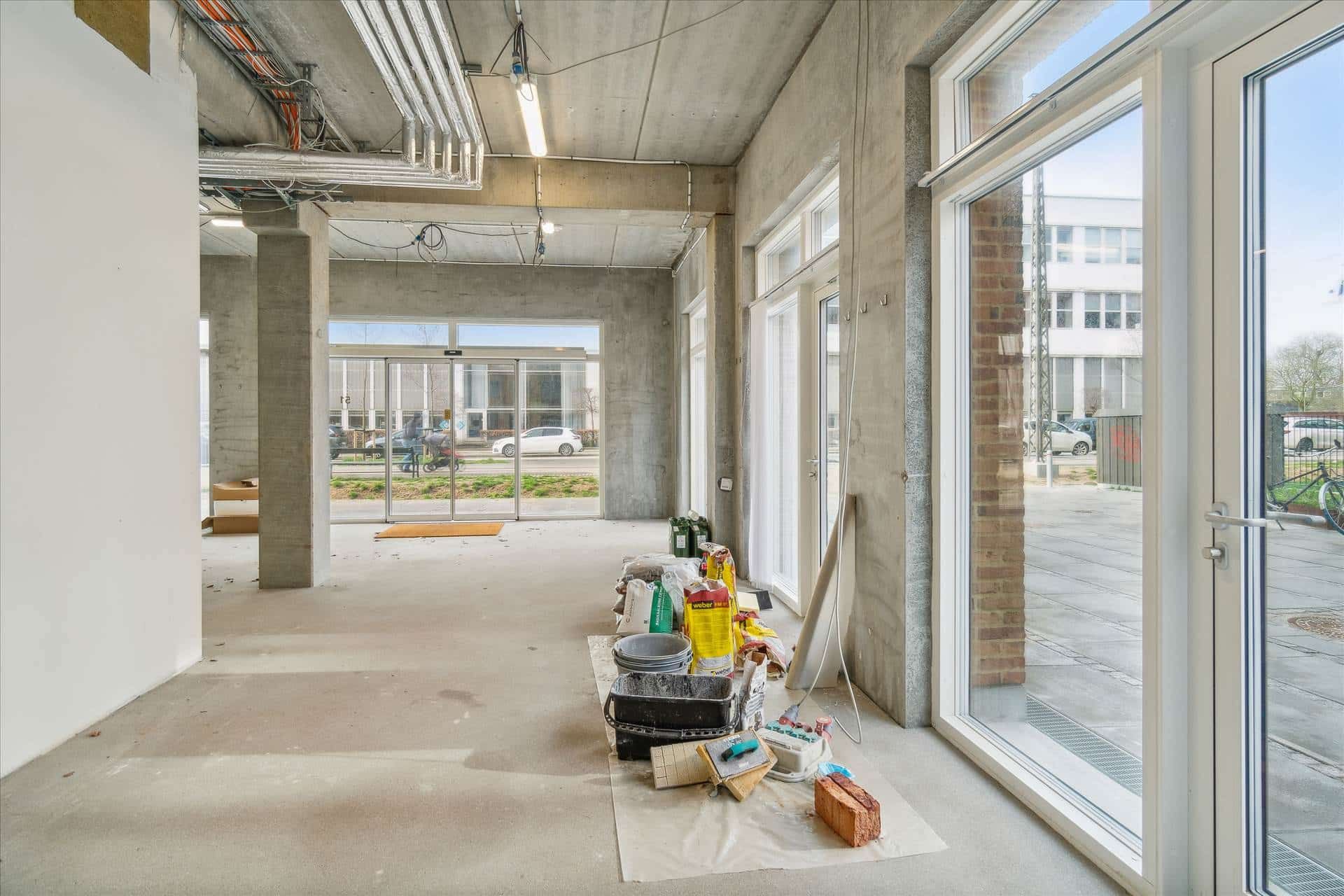 Et moderne kontorlokal med lyse vinduer og glasdøre. Forskellige byggematerialer og værktøj er spredt ud på gulvet. Udenfor