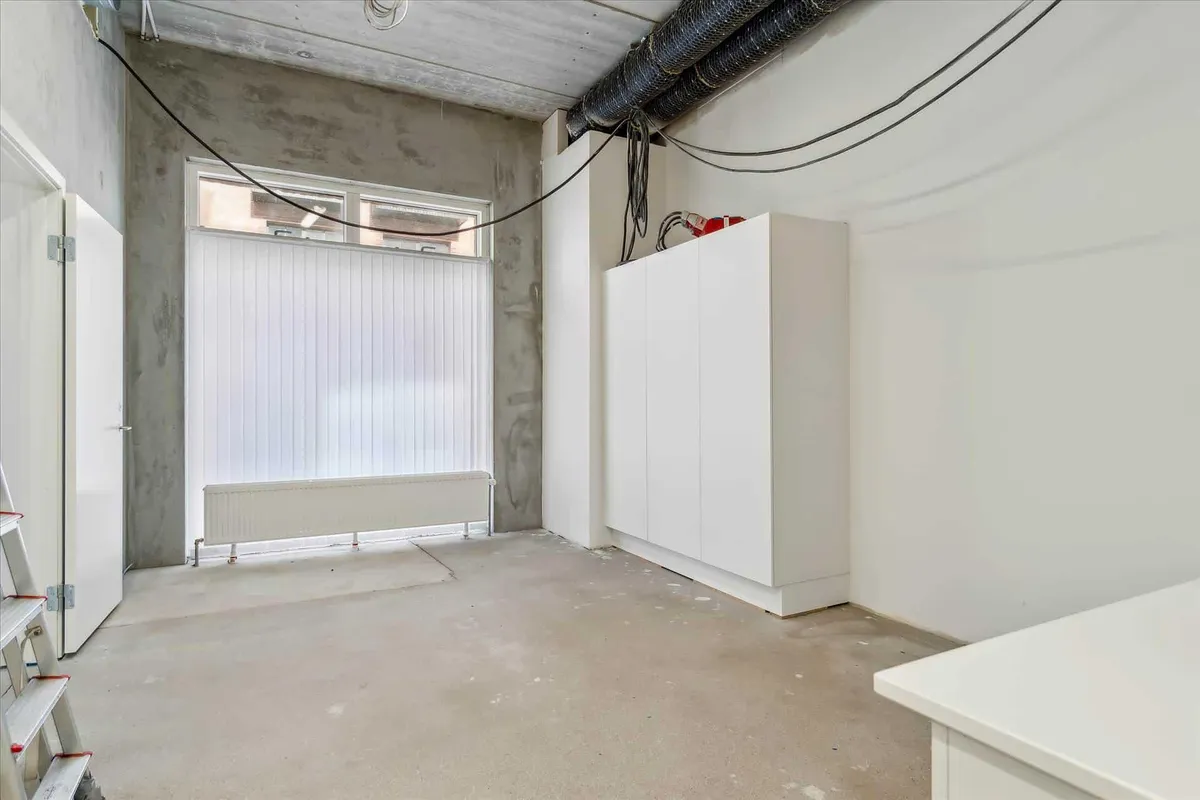 En tom værkstedslokal med hvidt væg, gulv og højt loft med kabel og rør. Der er en radiator og et vindue med jalousier og et